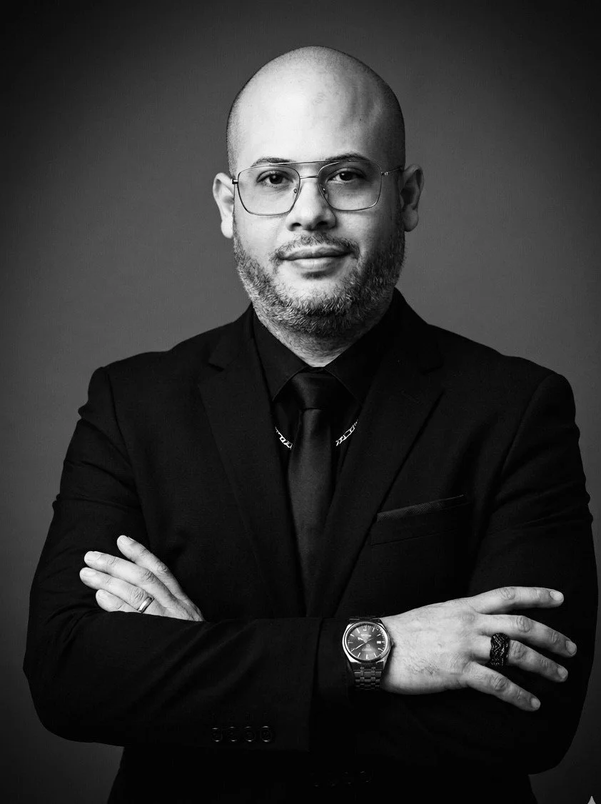 Black and white portrait of a man with shaved head, glasses, beard, wearing a dark suit, tie, watch, and rings, with arms crossed, against a plain background.