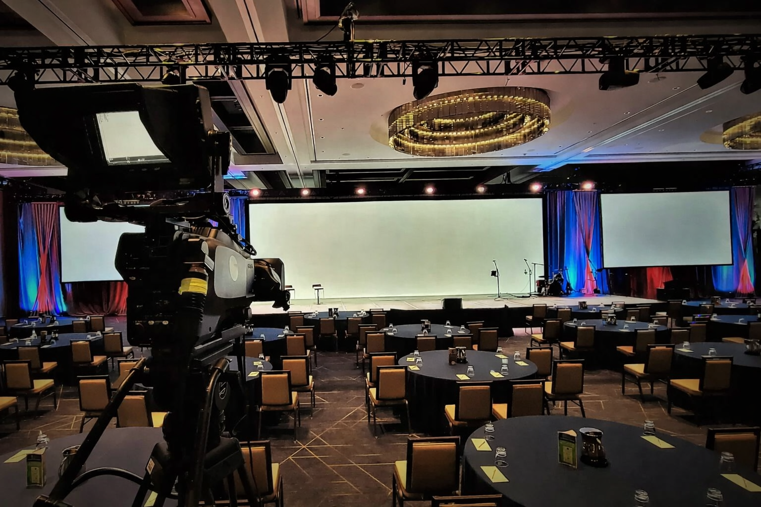 Camera pointed at an empty conference room with a stage, multiple large screens, and round tables arranged for speakers and attendees.