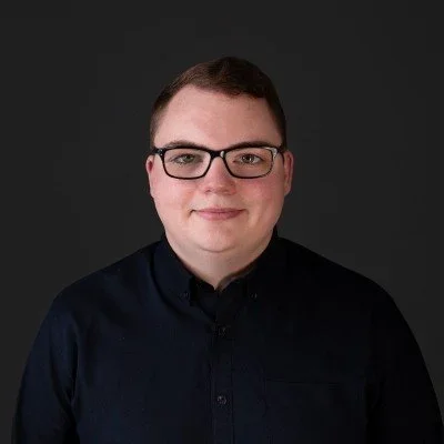 Portrait of a person with short dark hair, glasses, and a black shirt, smiling against a dark background.