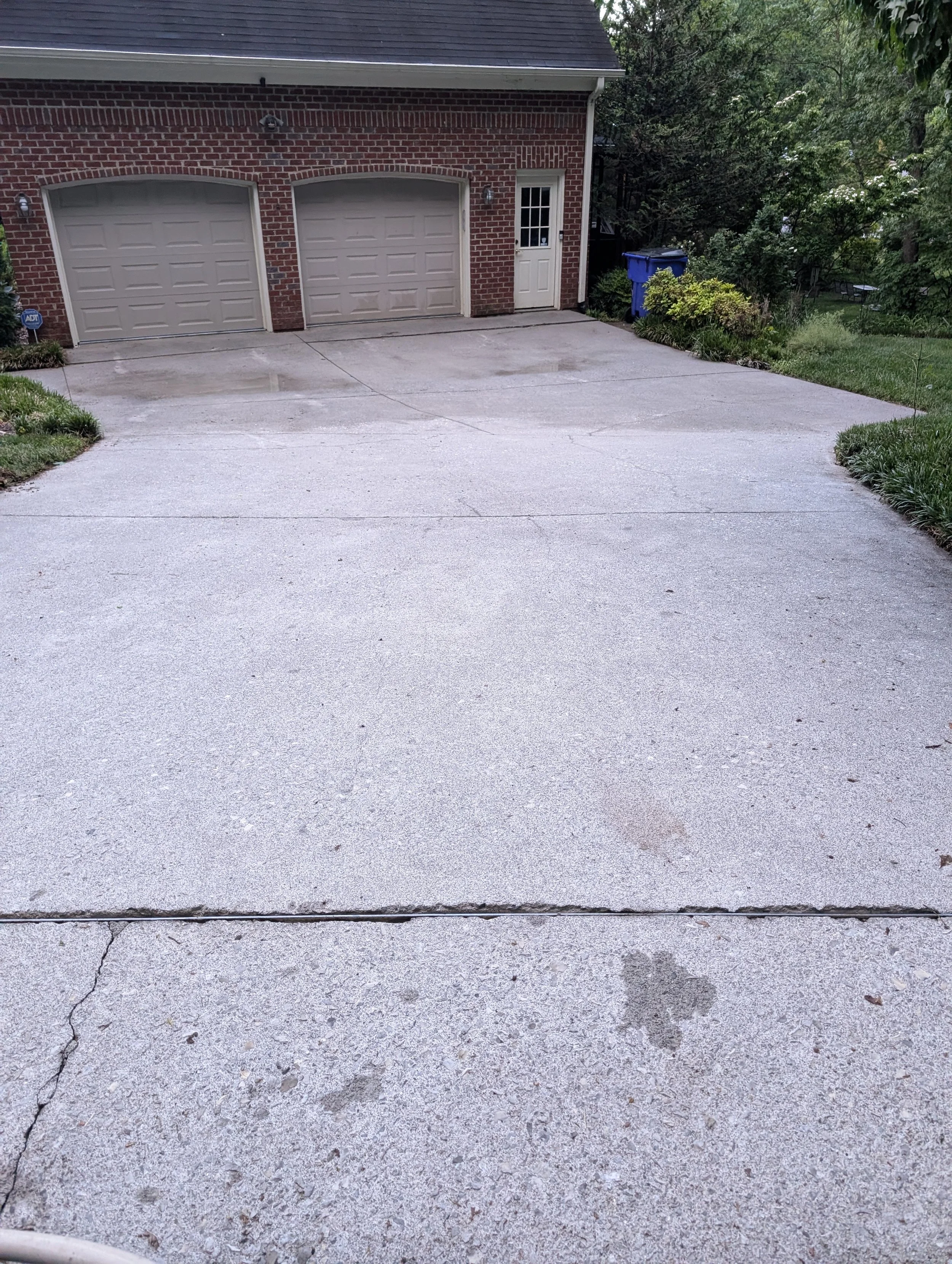 Clean concrete driveway leading up to a two-car garage with brick exterior and a side door. Green bushes and trees are on the right side, with a blue trash bin nearby.