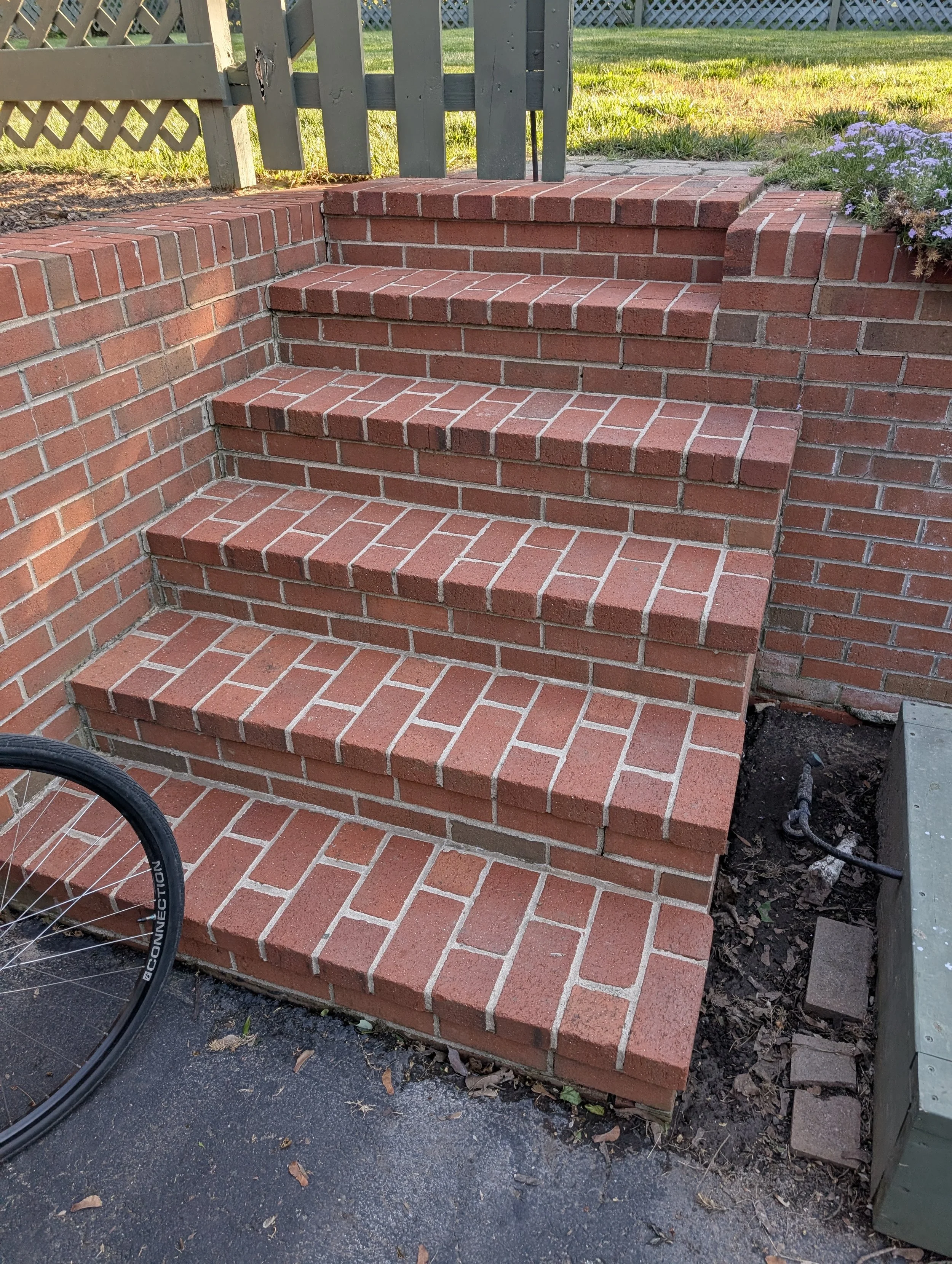 Freshly pressure washed brick stairs leading up to a small gate, with grass and purple flowers on the side, and a bicycle tire partially visible in the foreground.
