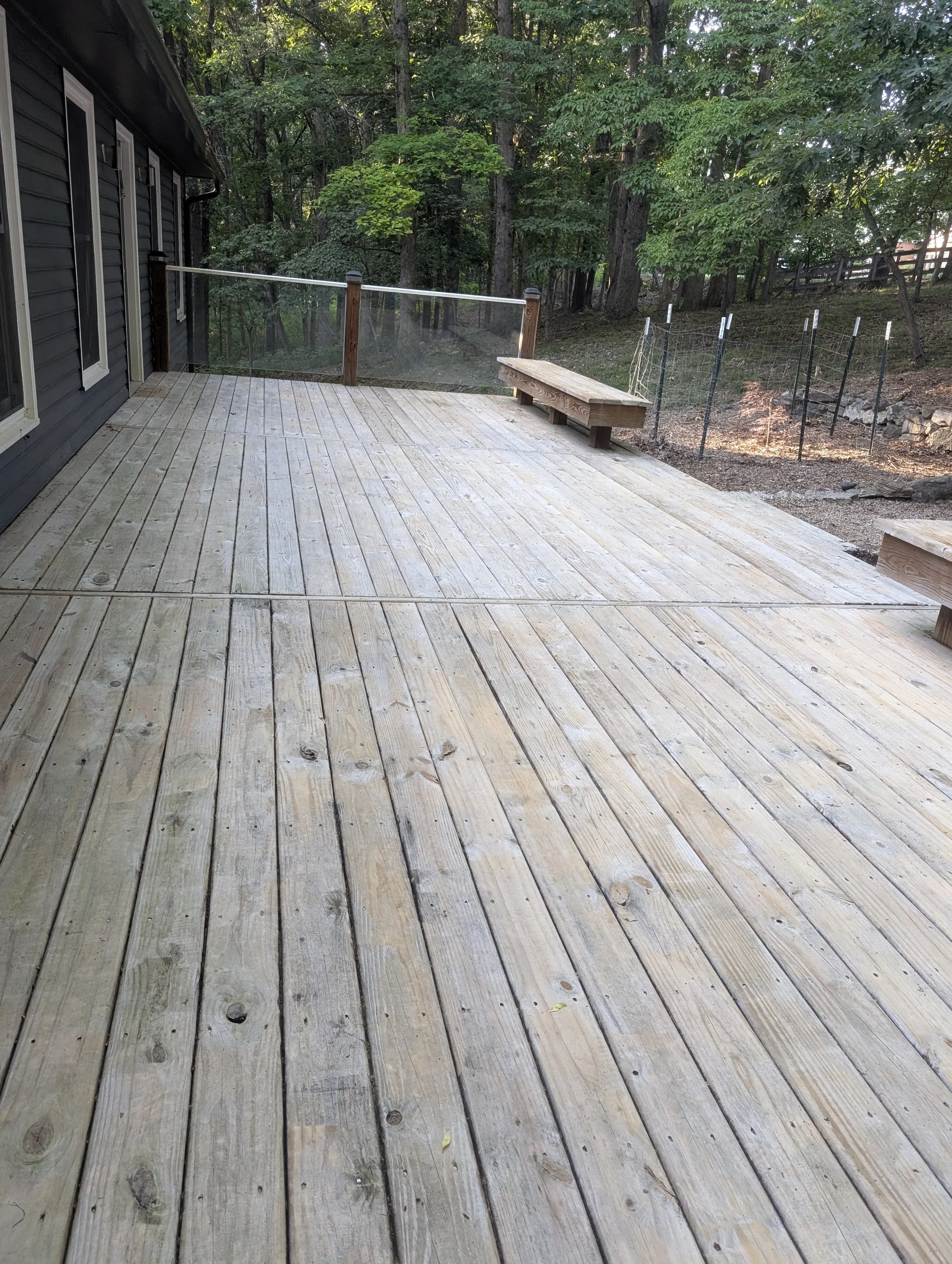 A wooden deck is attached to a house with black siding and white-trimmed windows. The deck has a glass railing with wooden posts and a built-in wooden bench. The background features a wooded area with trees and a dirt yard.