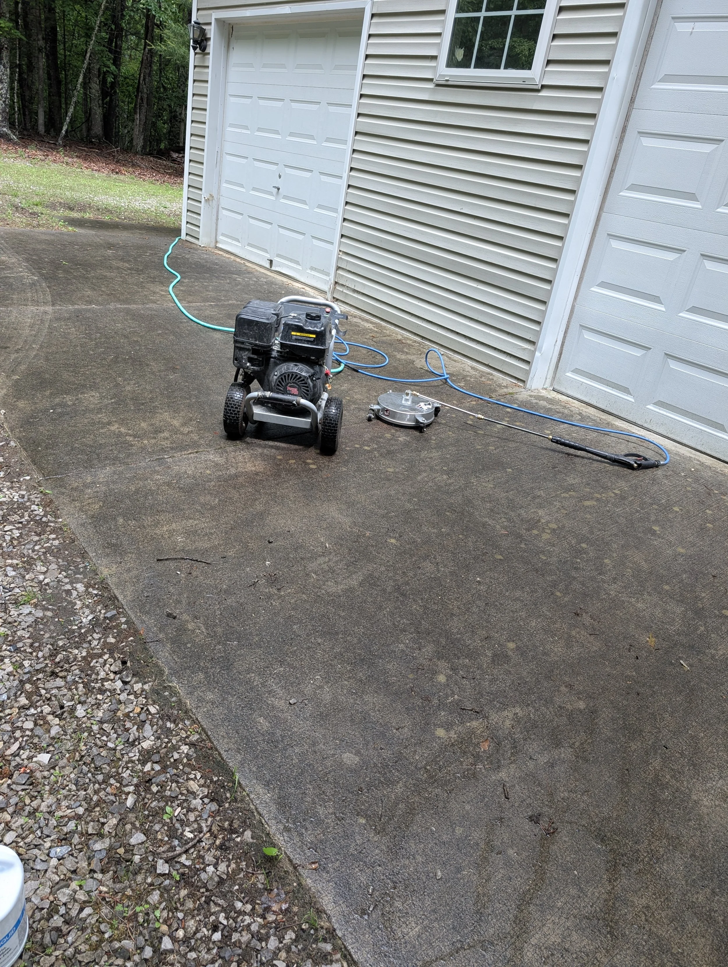 A pressure washer with a black engine and wheels, connected to a long wand, sitting on a dirty concrete driveway next to a white garage door and beige horizontal siding, with a wooded area in the background.
