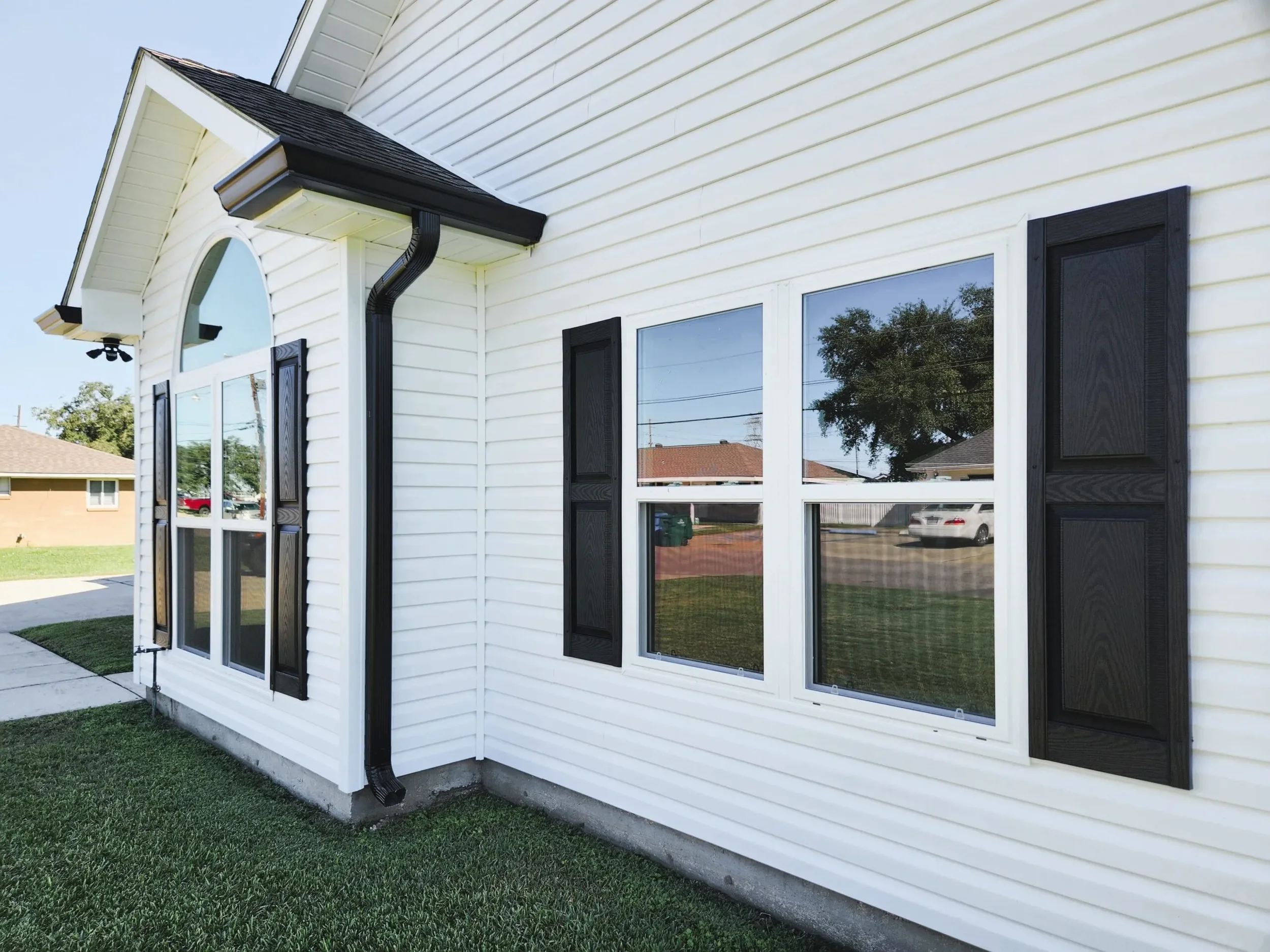Close-up of a white house exterior with black window shutters, a black gutter downspout, and windows reflecting trees and neighboring houses, with a small grassy area in the foreground.