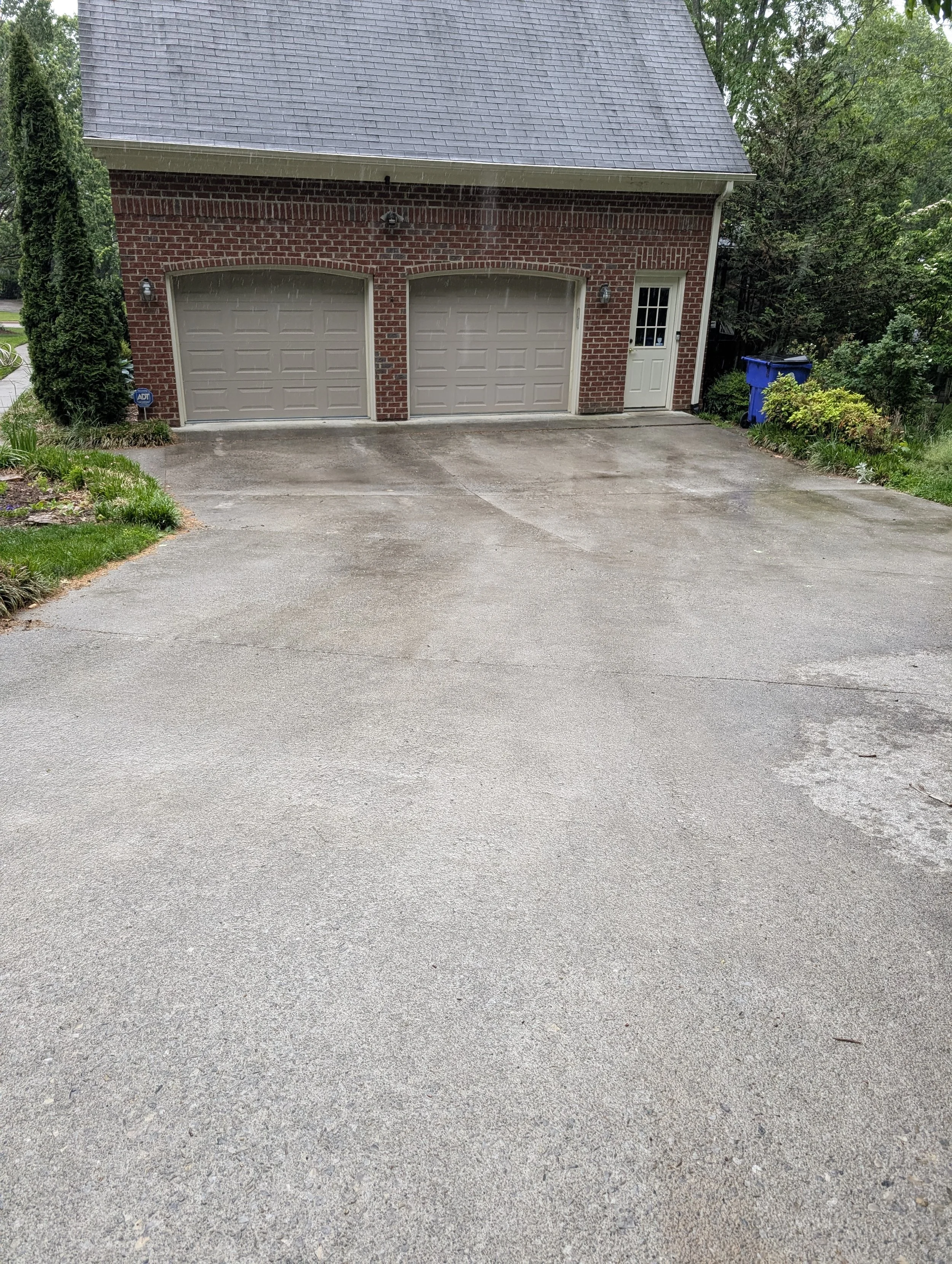 A driveway in front of a brick garage with a door and two garage doors. There is greenery and a blue recycling bin to the right.
