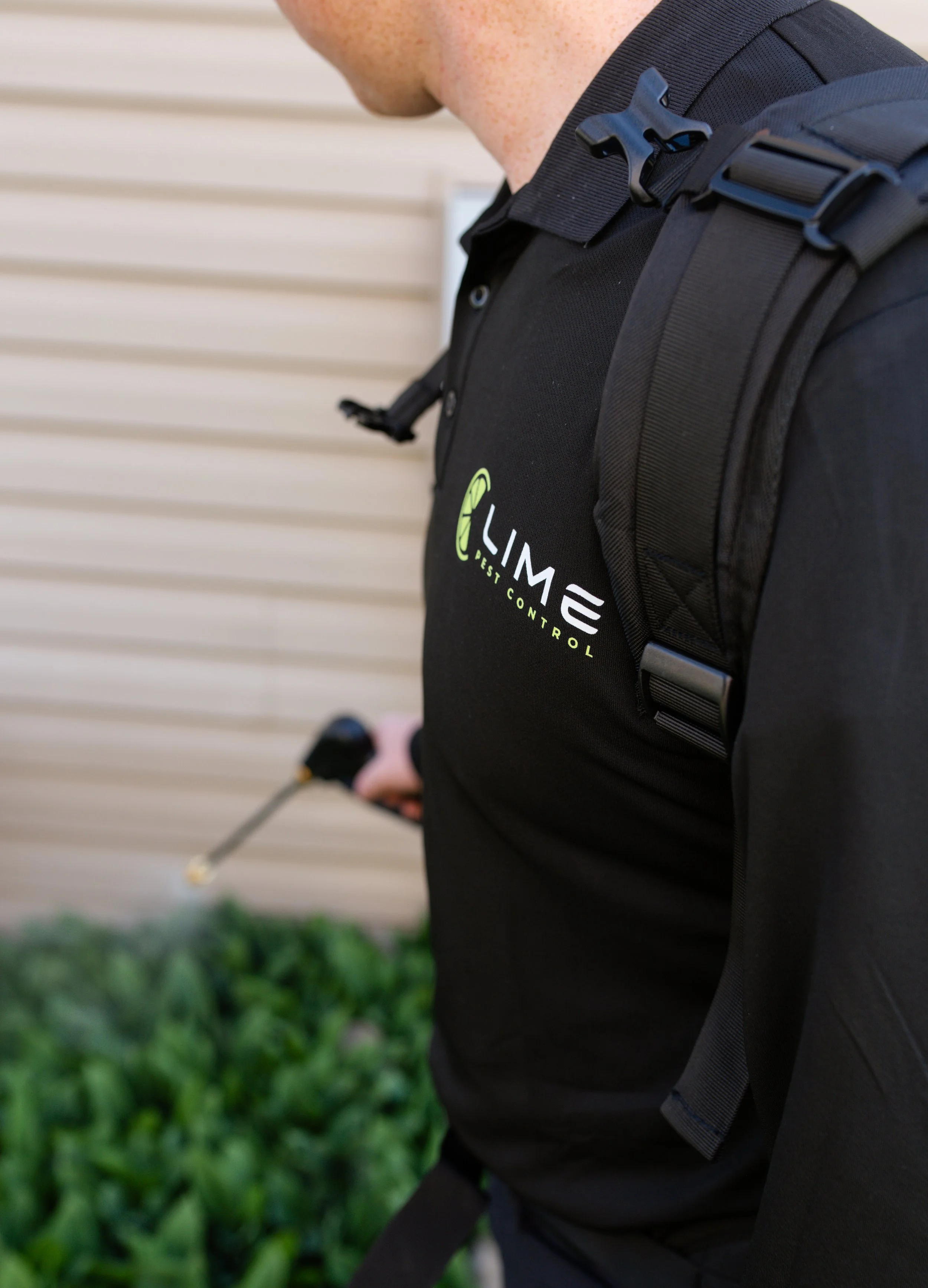 Close-up of a person wearing a black shirt and a black backpack, holding a golf club, standing outdoors near greenery and a beige siding wall.