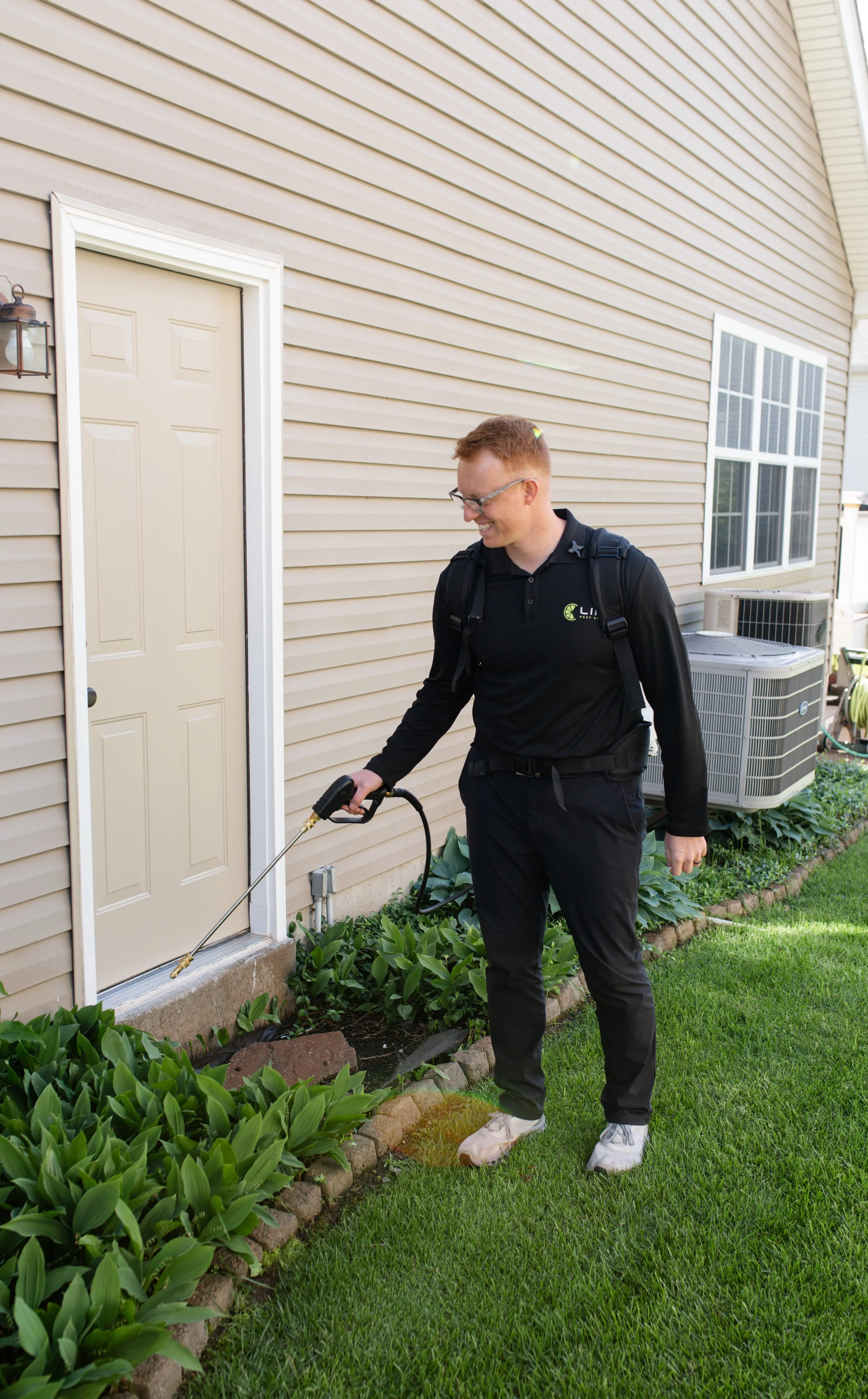 A man with glasses, red hair, wearing a black long-sleeve shirt, black pants, and white sneakers is using a weed spray tool to treat plants along the side of a beige house. He is smiling and carrying a backpack-style sprayer. The house has beige siding, a white door, and a window with grid panes. There is a small garden bed with green plants and bricks, and an air conditioning unit is visible in the background.