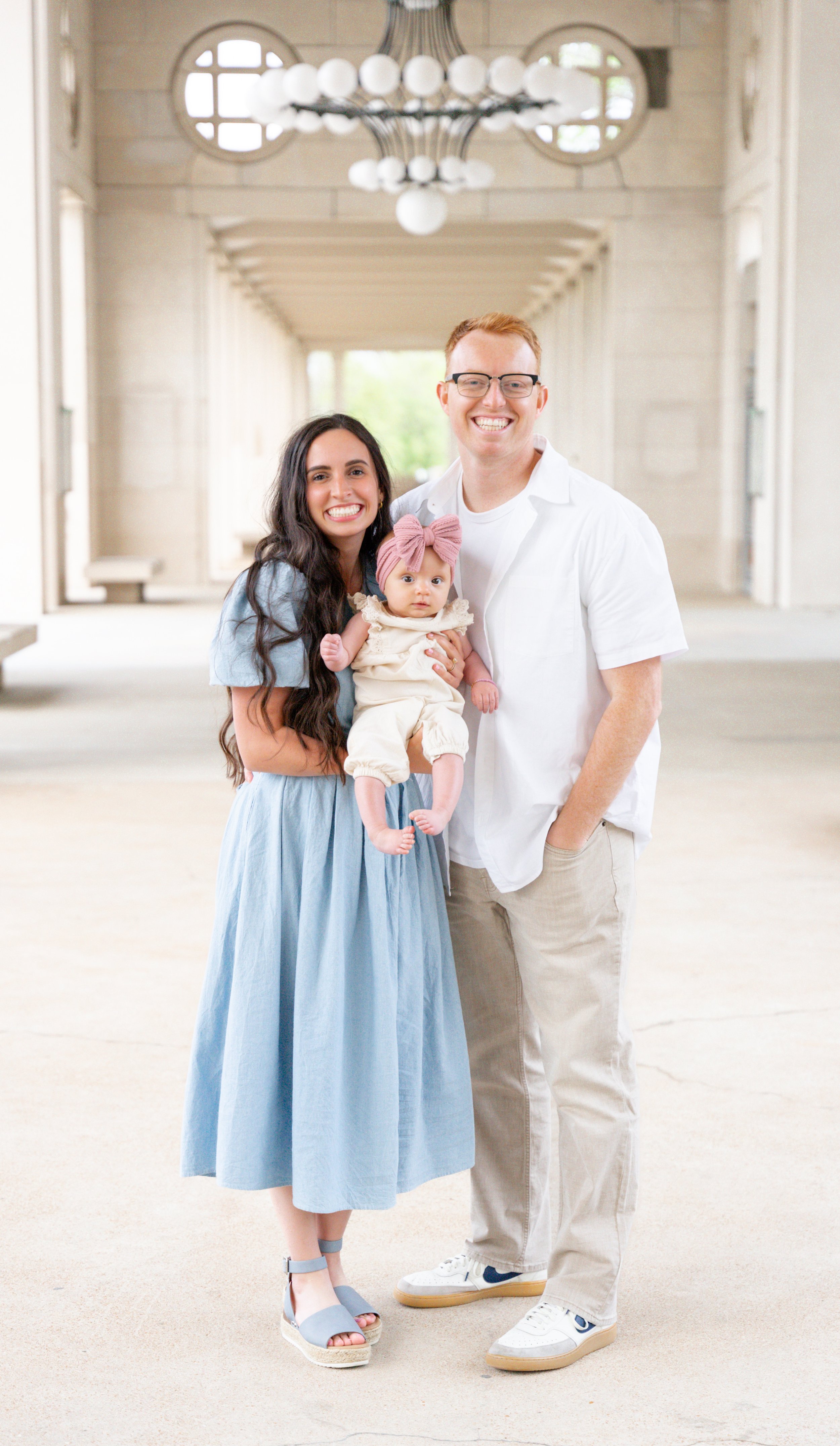A family of three standing in a bright, spacious corridor with modern lighting and circular window details, smiling at the camera. The woman wears a blue dress and high heels, the man is in a white shirt and sneakers, and the baby girl has a pink headband and beige outfit.