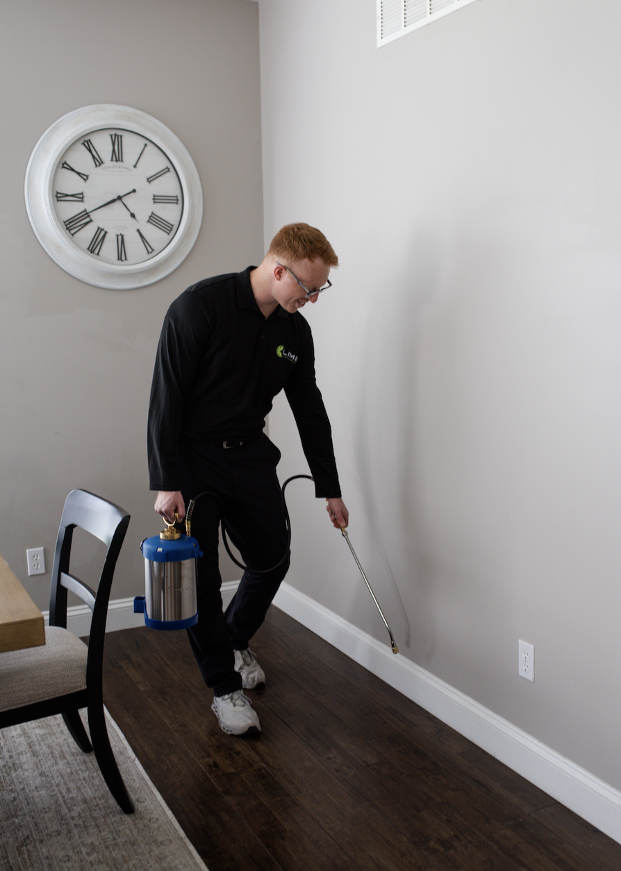 A pest control technician in black uniform spraying pesticide along a wall in a residential dining room with a large wall clock and dark wood floor.