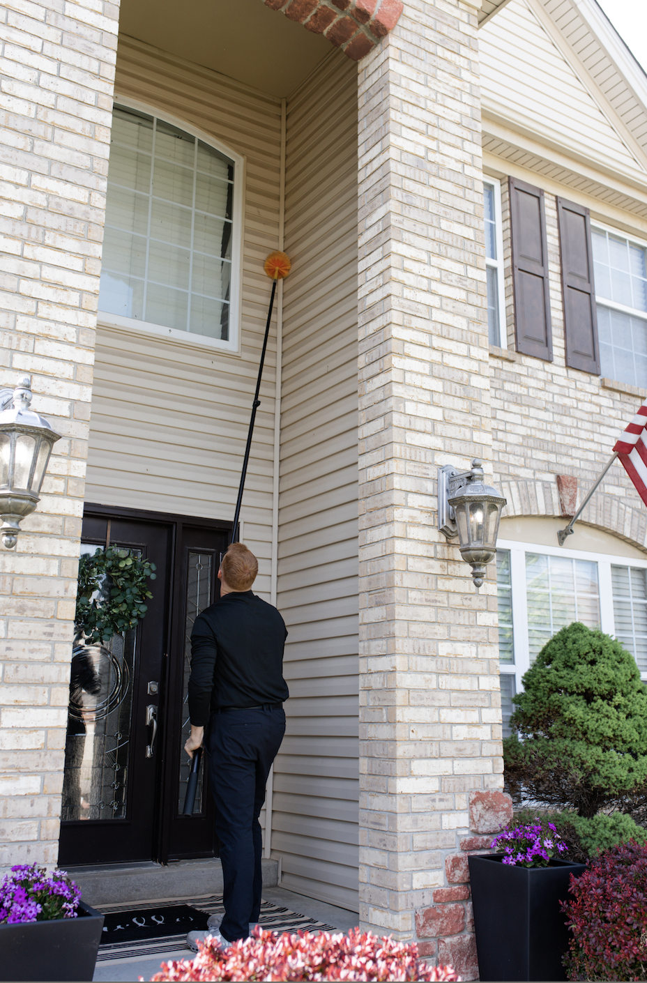 A man using a telescoping duster to clean the upper corner of a house's exterior wall near a window.
