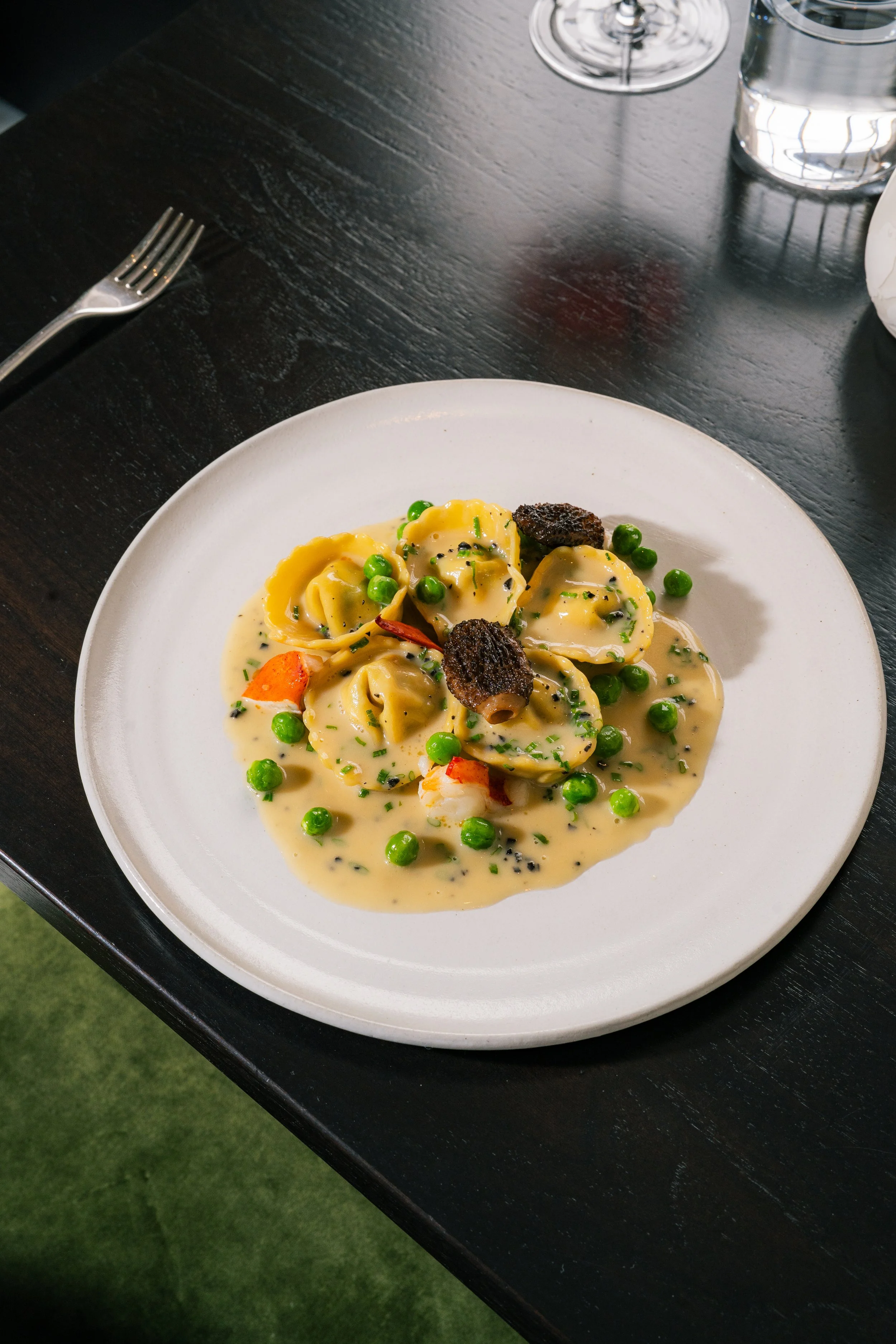 Plate of tortellini pasta with a creamy sauce, green peas, and black pepper, garnished with black truffle slices and chopped herbs, on a white round plate at a dark wooden table with a fork, a glass of wine, and a glass of water.