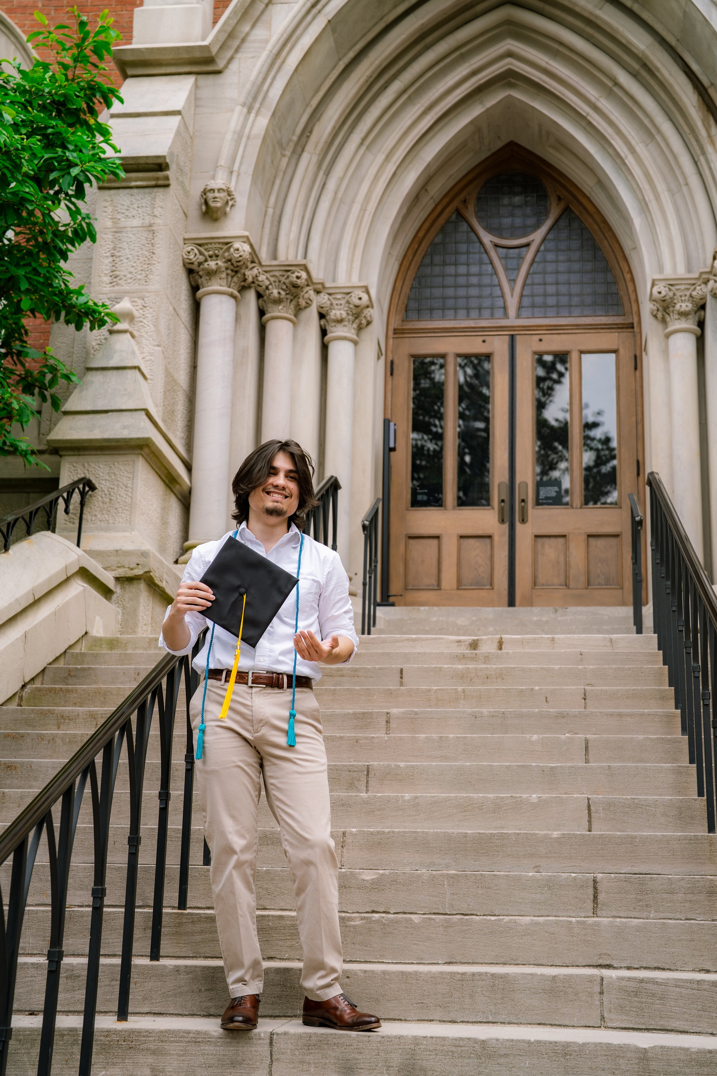 A young man with shoulder-length brown hair, beard, and smile, standing on stone steps outside a historic building with arched doorway. He is holding a graduation cap and a diploma, wearing beige pants, a white shirt, and brown shoes, with a blue tas