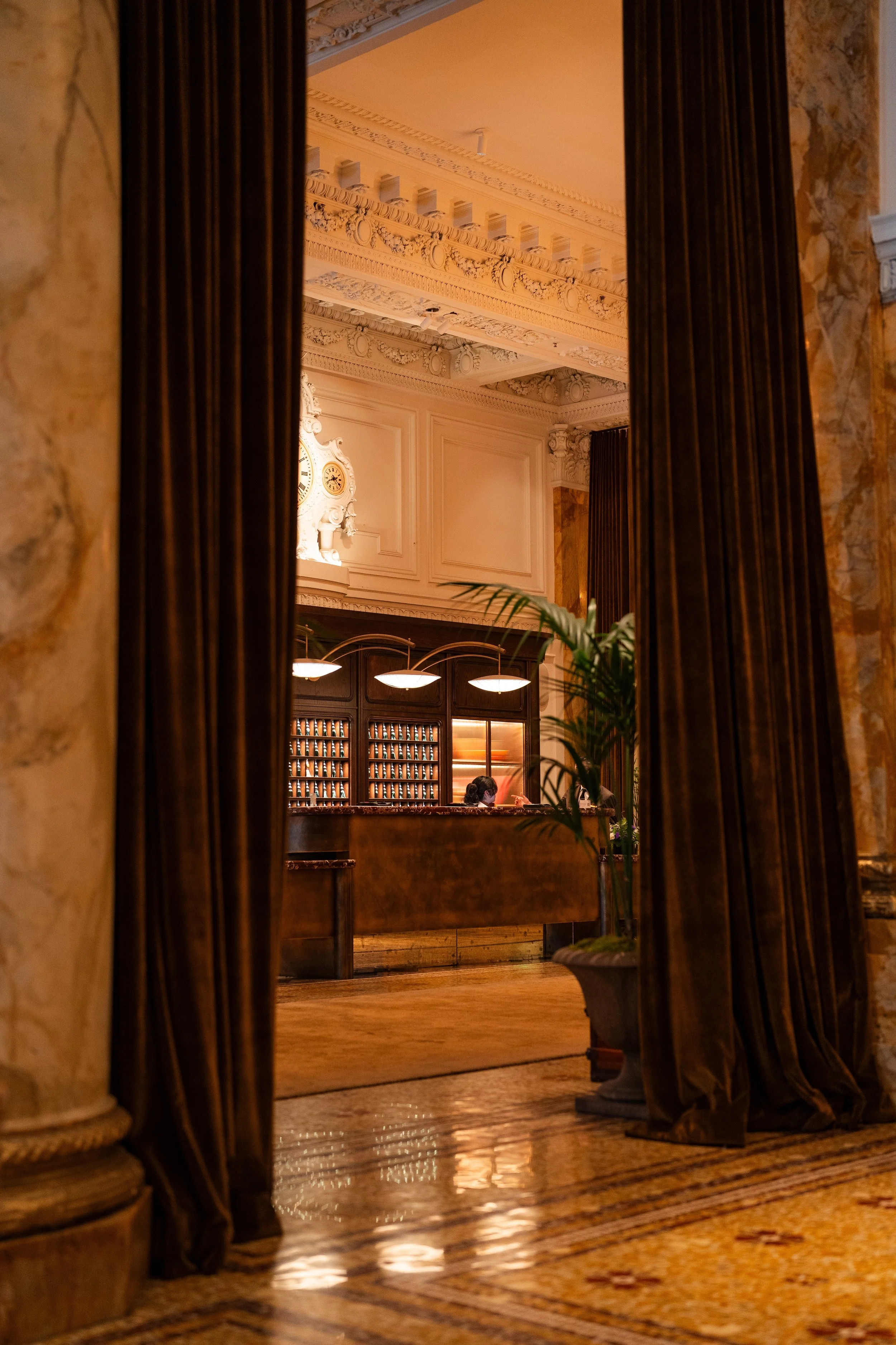 Interior of a luxurious building with ornate walls, a decorative clock, wooden furniture, and warm lighting, viewed through curtains.