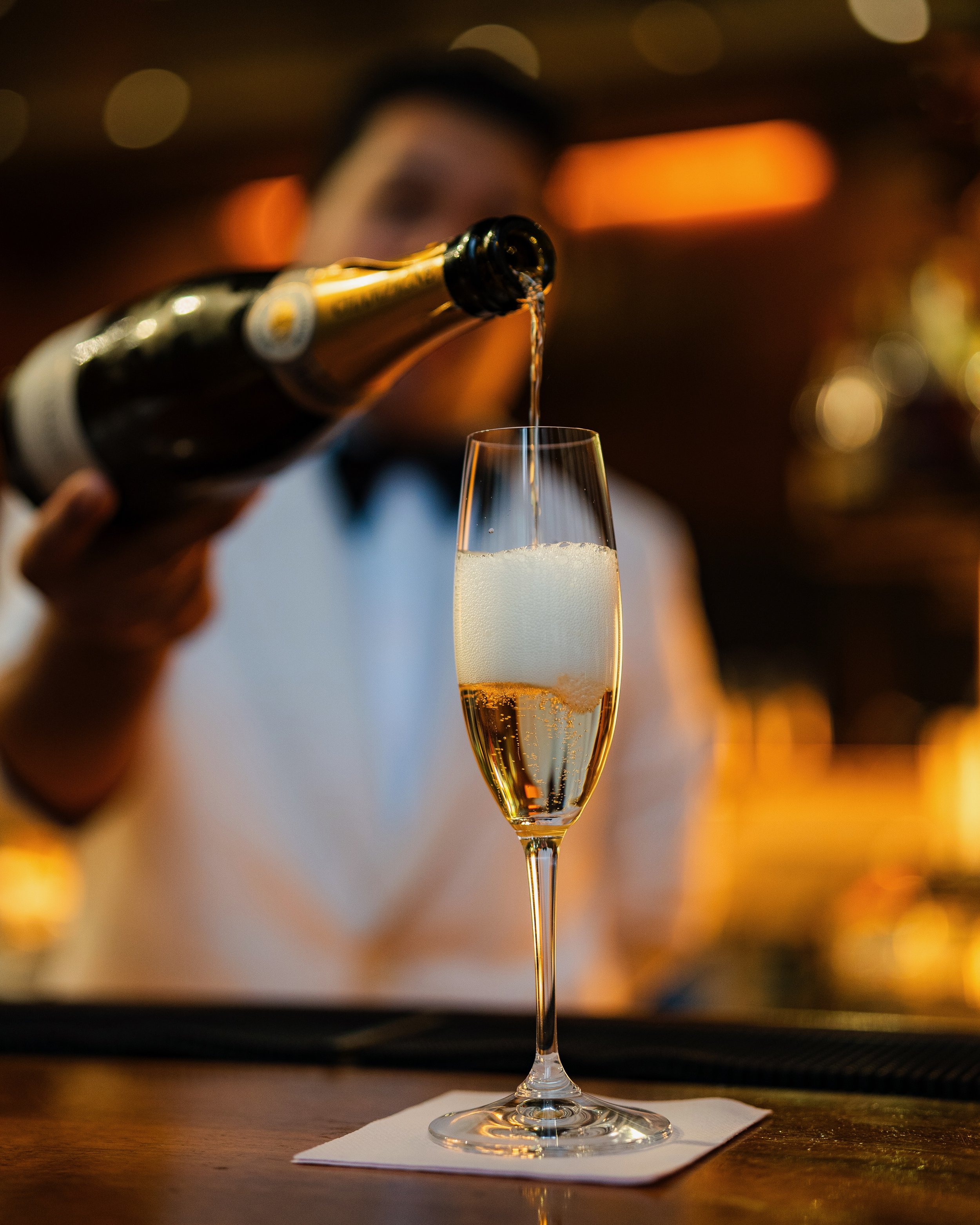 A person is pouring champagne into a flute glass, which is placed on a white napkin on a wooden table, with warm, blurry lights in the background.