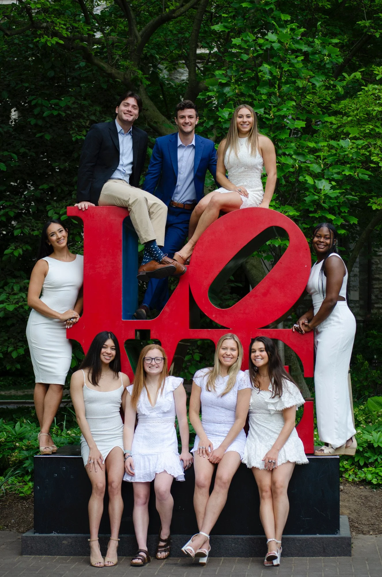 Group of young adults posing in front of a large red 'LOVE' sign outdoors with greenery background.