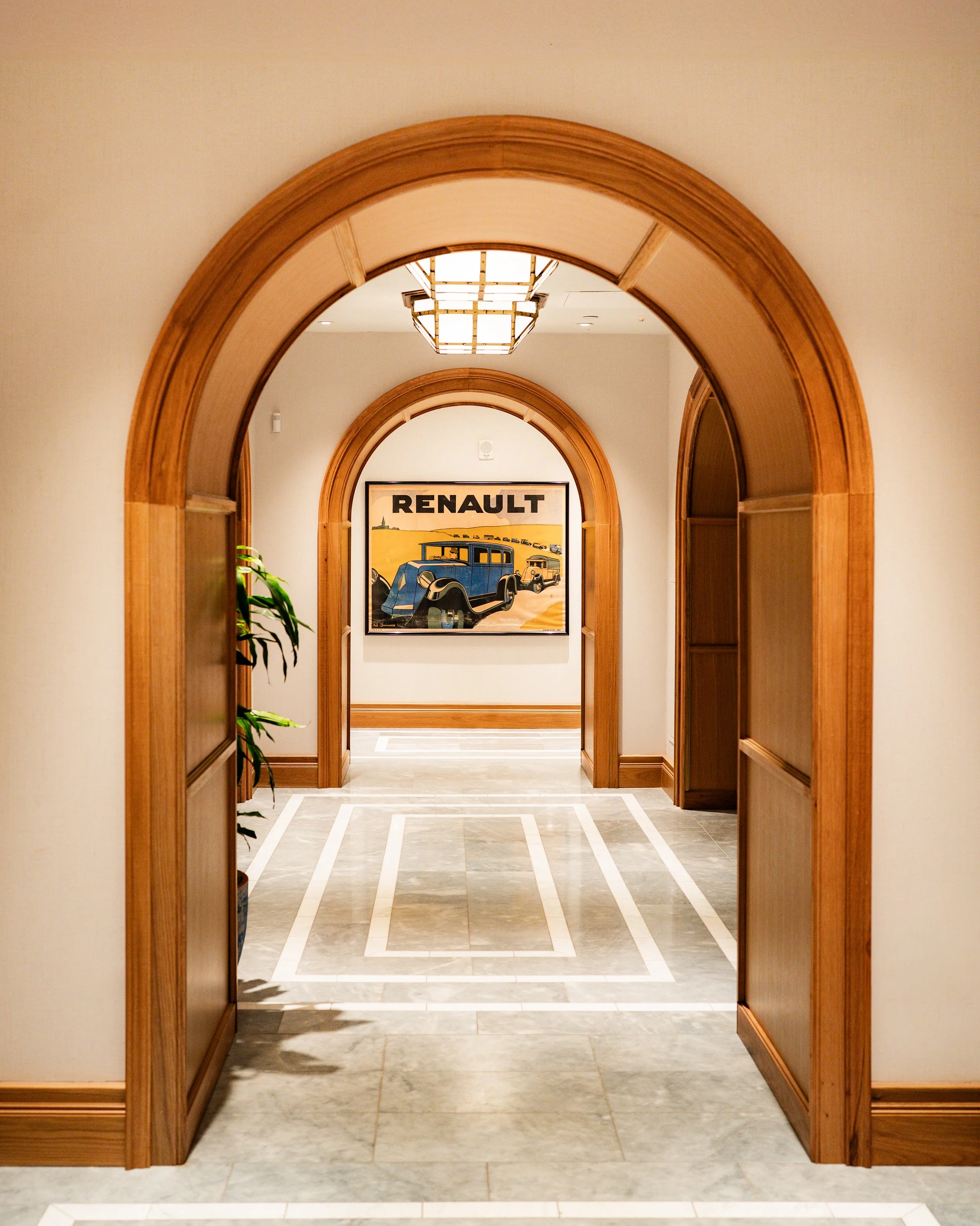 A hallway with wooden archways and a framed vintage Renault car poster on the wall. There is a ceiling light fixture and potted plant on the left.