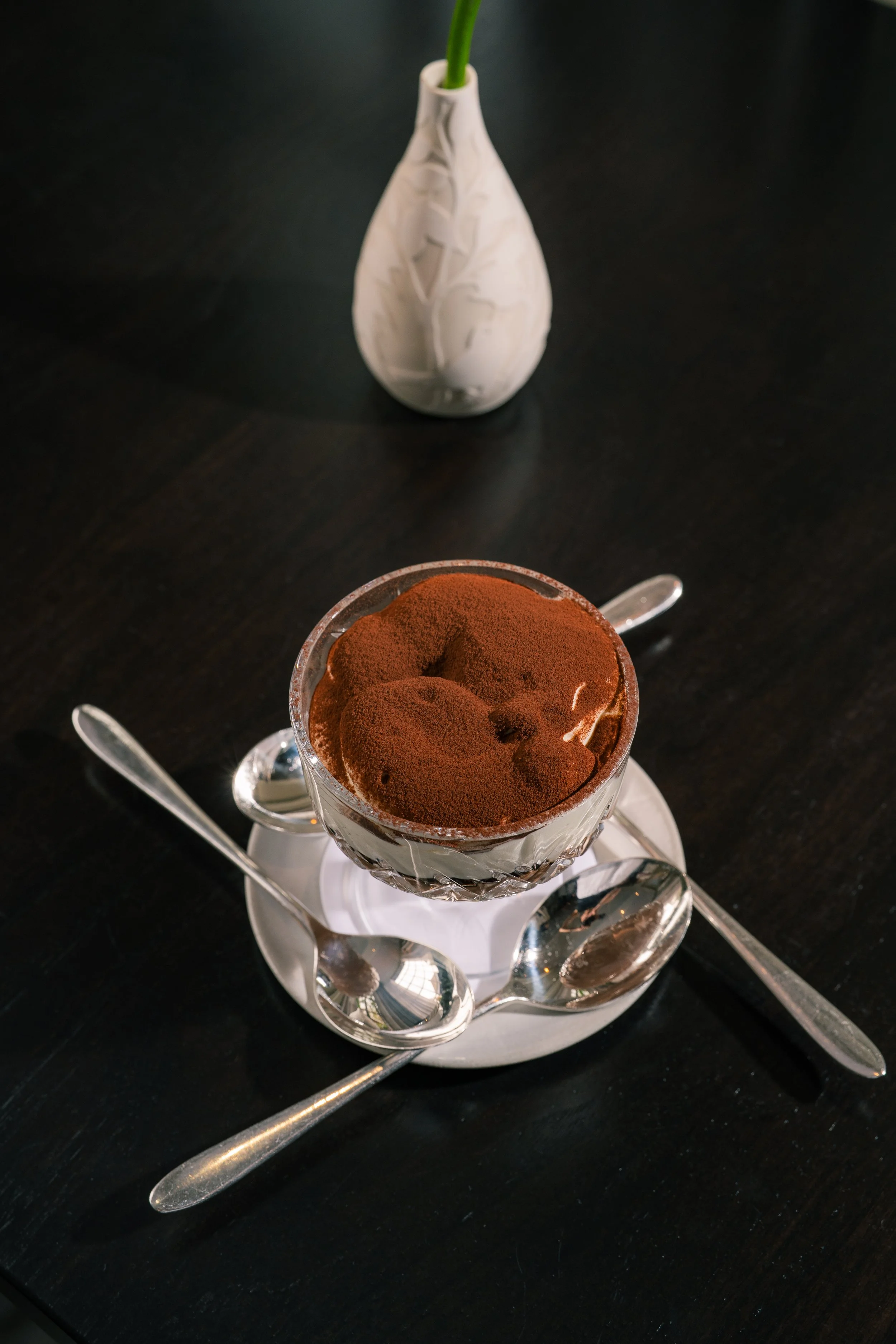A glass cup of tiramisu with cocoa powder on top, placed on a silver tray with spoons, on a black table, and a white vase with a green stem in the background.