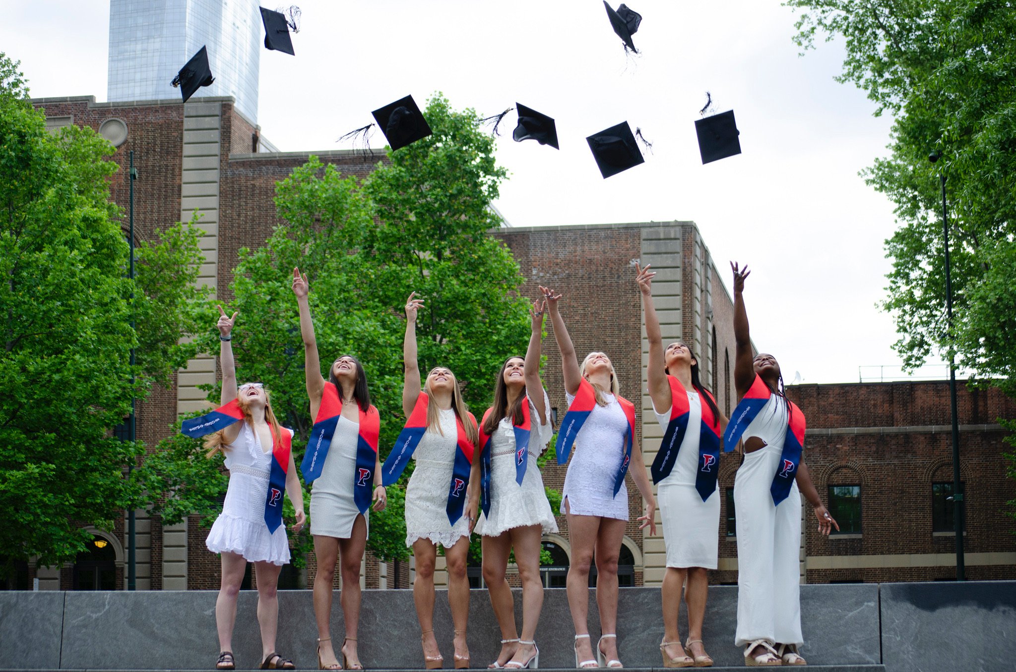 A group of women in white dresses and stoles celebrating graduation outdoors as they throw caps into the air against a backdrop of trees and brick buildings.