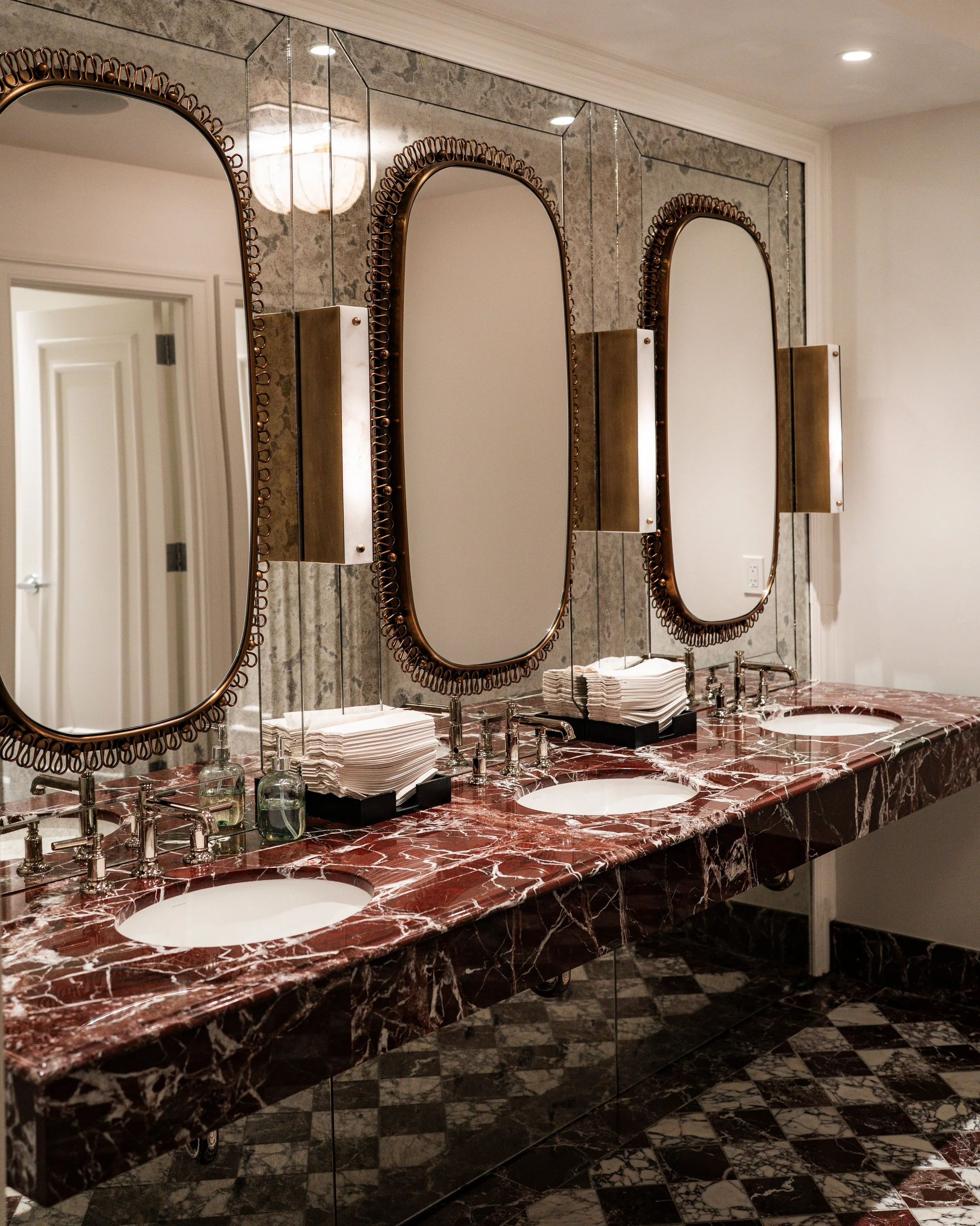 A red marble countertop with three white sinks, each with a silver faucet and a soap dispenser. Above the sinks are two rectangular wall-mounted mirrors with decorative bronze frames. Towels are stacked on the counter with reading glasses and a black