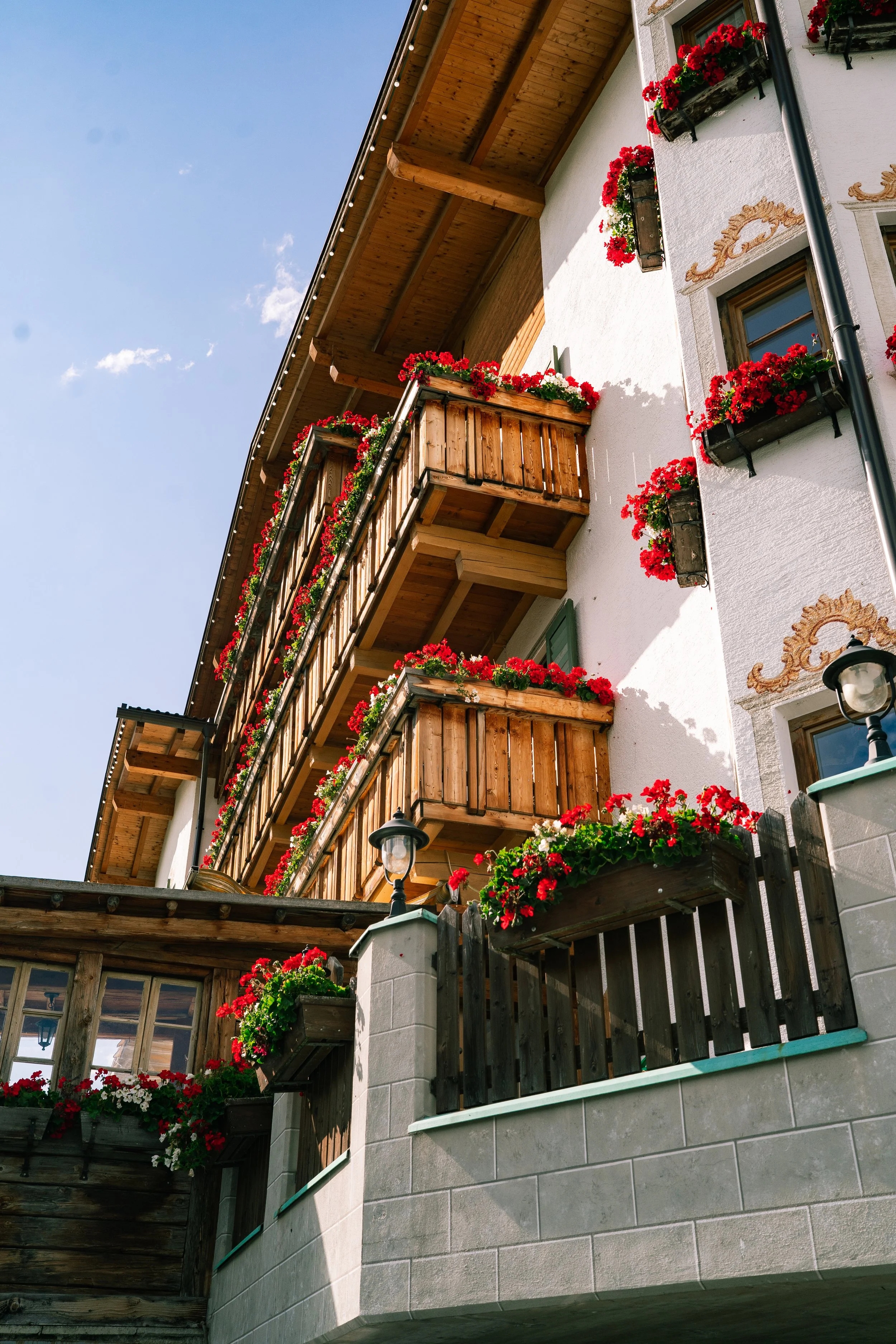 A multi-story building with wooden balconies decorated with red and white flowers in window boxes. The building has a white exterior with decorative paint details around the windows and a sloped roof with under the eaves lighting. The sky is clear an