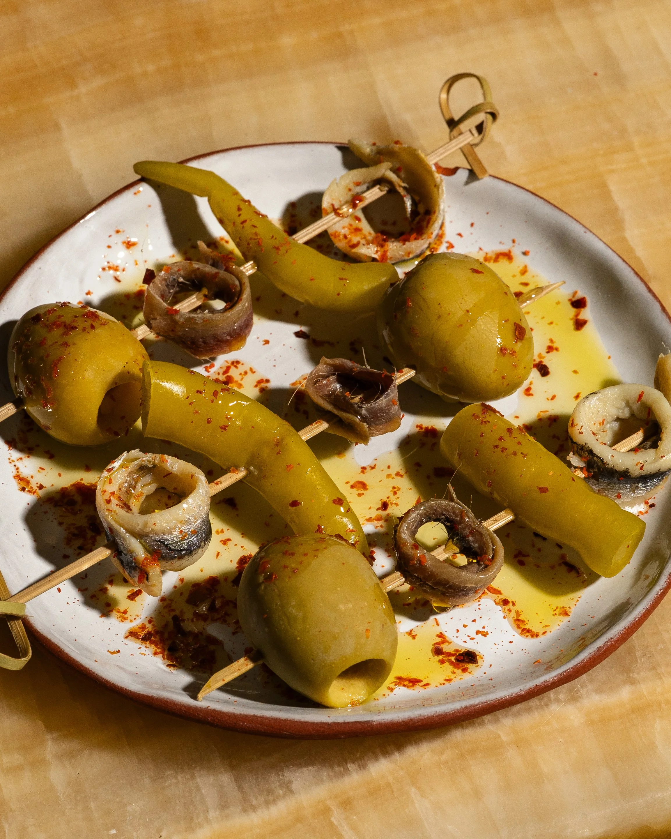 Skewers with green olives and chopped anchovies on a white plate garnished with red chili flakes, set on a wooden surface.