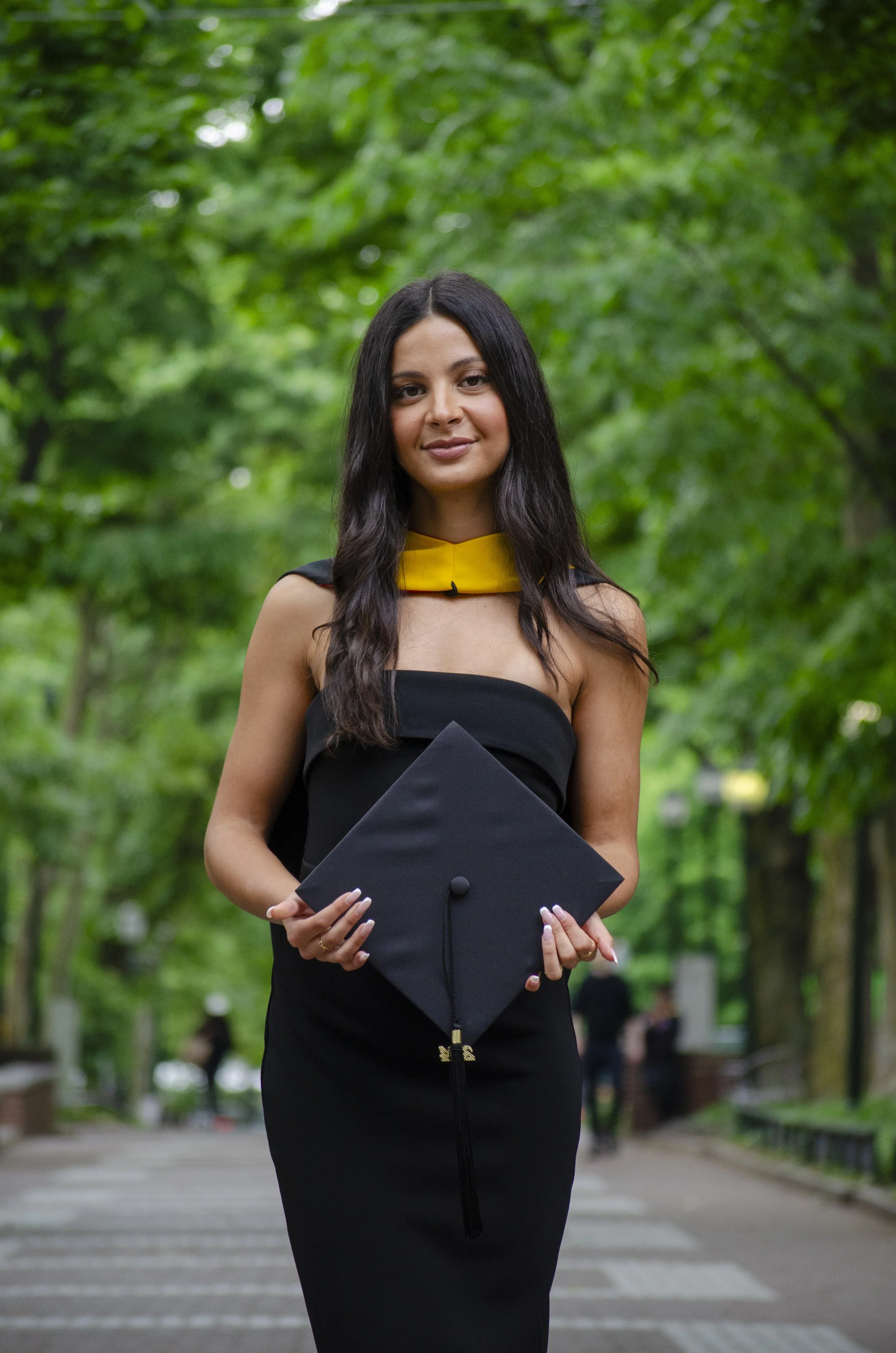 A young woman in a black dress holding a graduation cap outdoors in a park with green trees in the background.