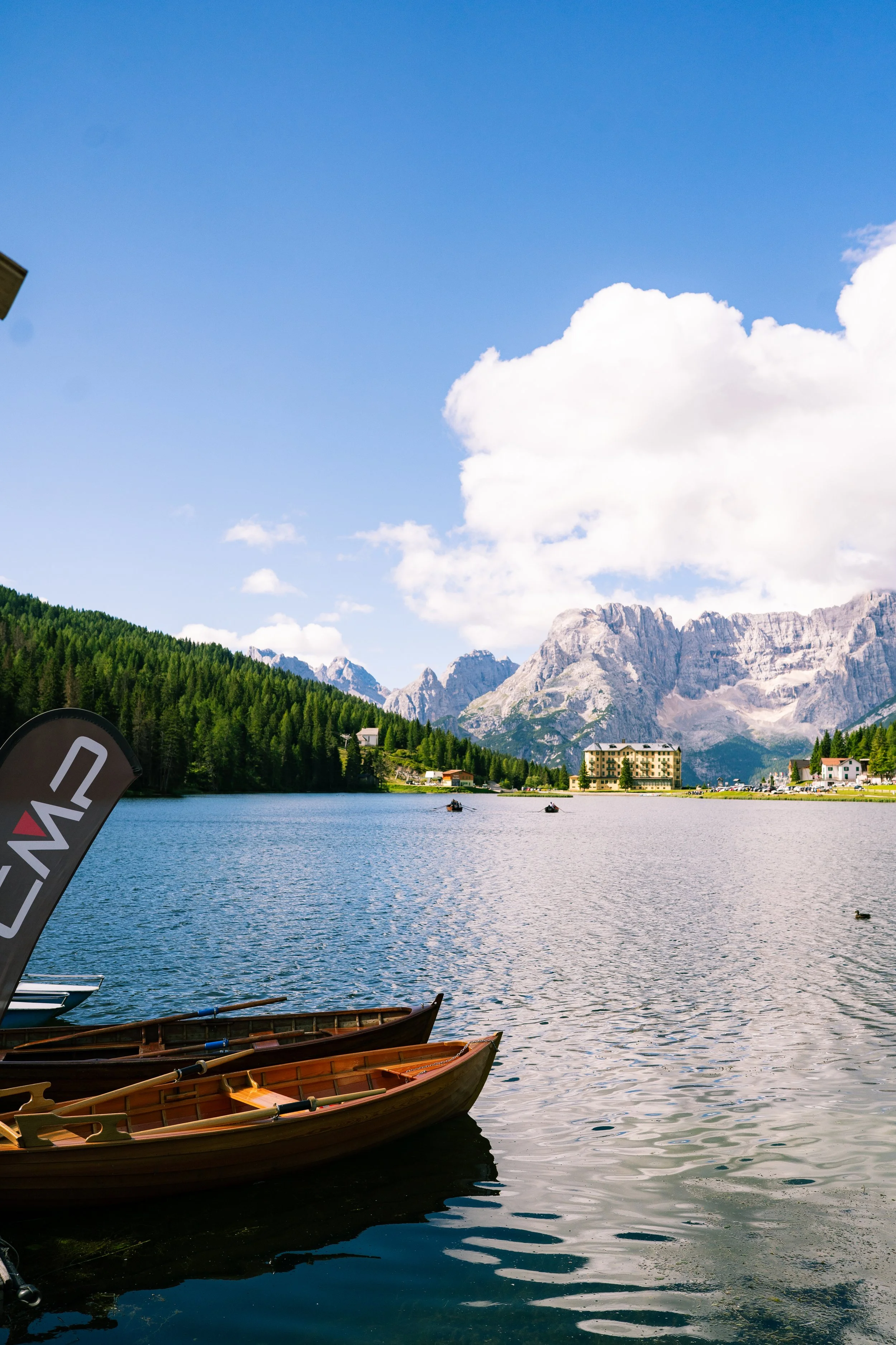 A peaceful lake with boats, surrounded by green forests, mountains, and buildings under a bright blue sky with white clouds.