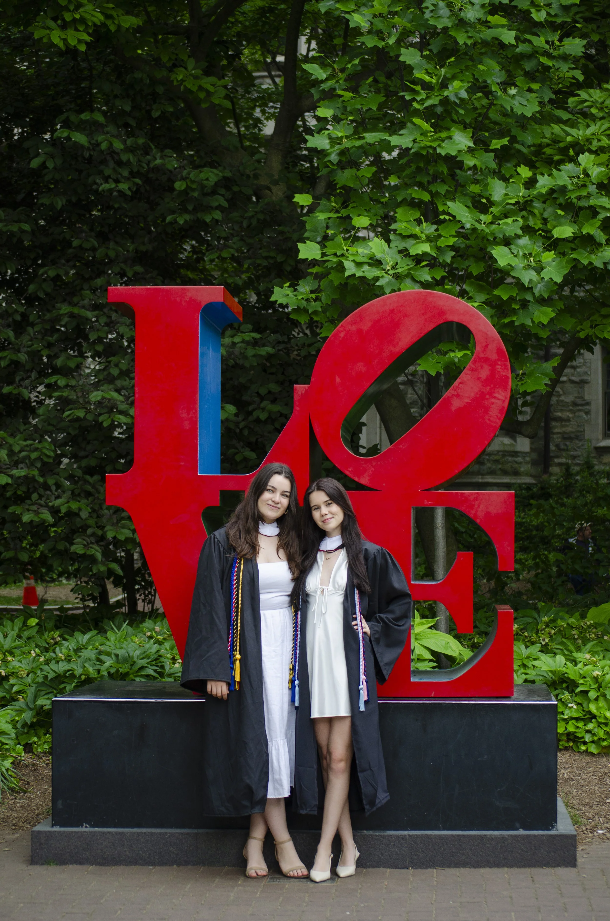 Two young women in graduation gowns and caps standing in front of a large red LOVE sculpture outdoors by green trees. They are smiling and posing for the photo.