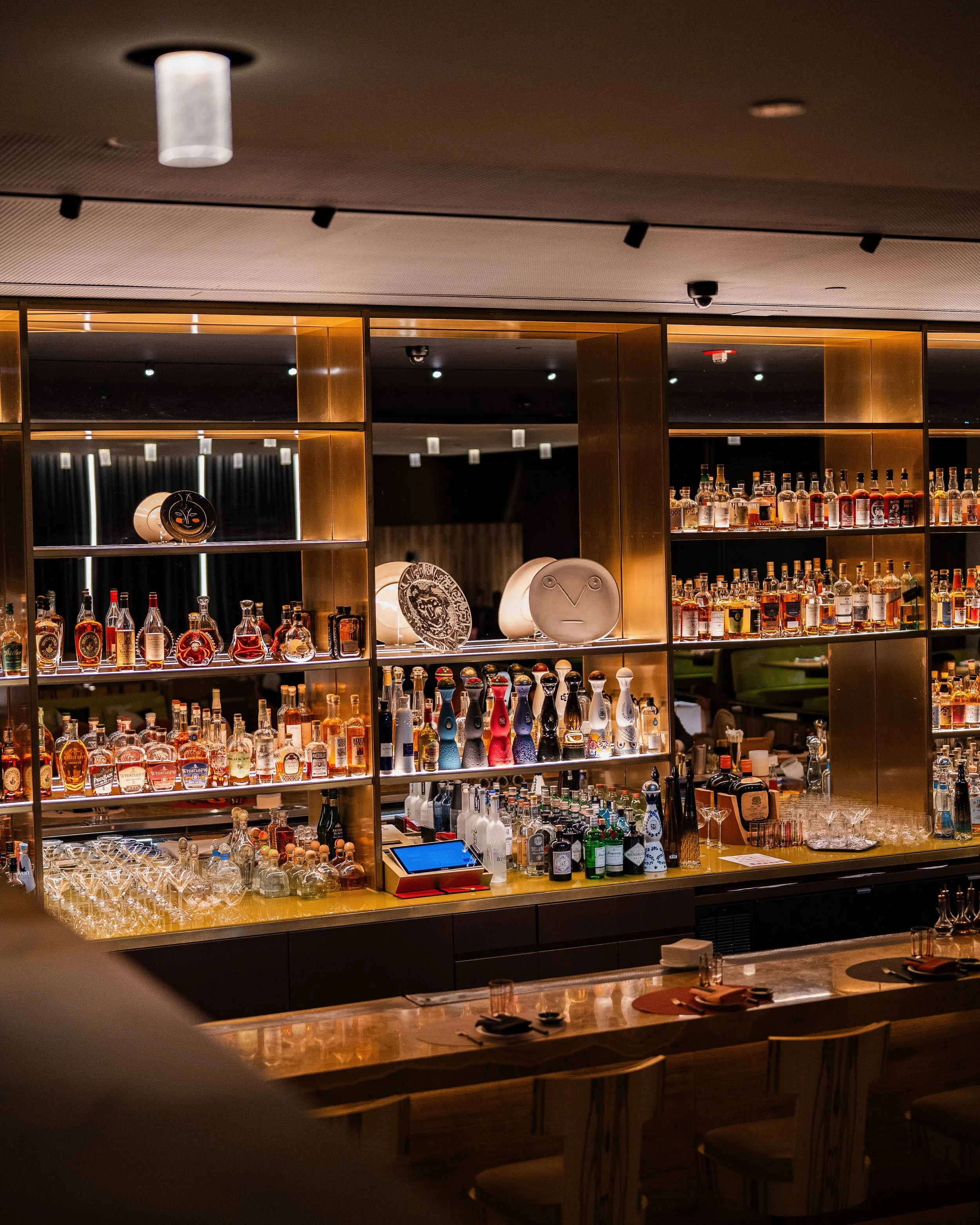 Interior view of a bar with shelves filled with various bottles of alcohol, illuminated with warm lighting. The bar counter has bar stools and is set with placemats, glasses, and utensils.