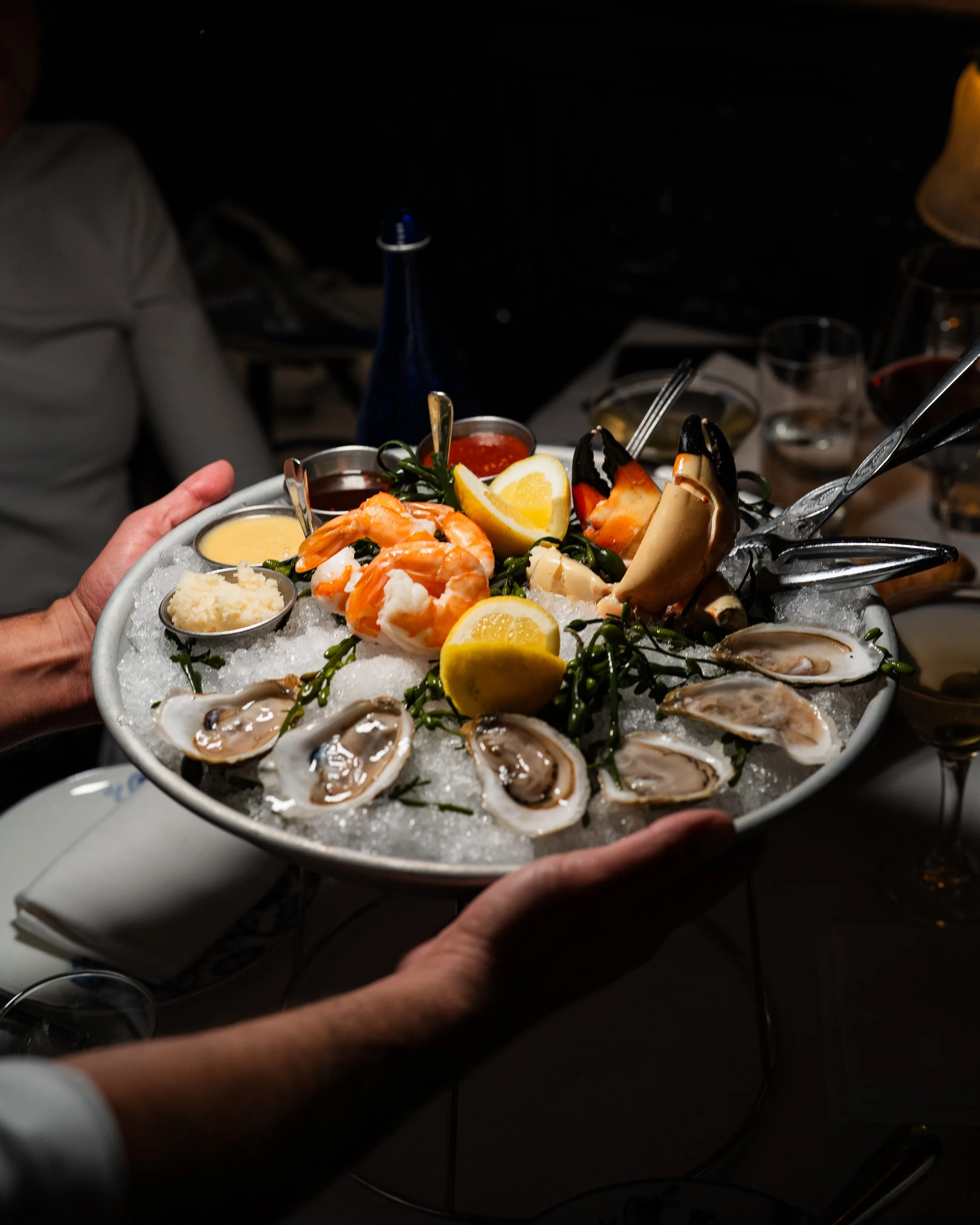 A seafood platter with oysters, shrimp, lobster tail, lemon wedges, and dipping sauces, served on a bed of ice, in a dimly lit restaurant setting.