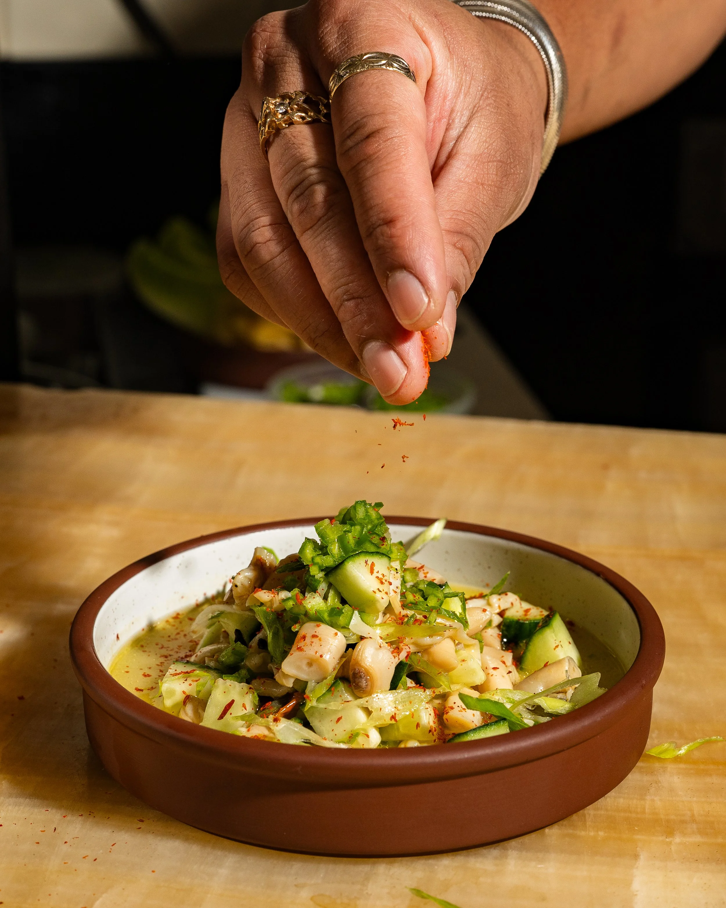 A person sprinkling red chili powder onto a bowl of vegetable salad. The salad contains chopped cucumbers, onions, and other vegetables. The person is wearing rings and a bracelet, and the scene is set in a kitchen or dining area.