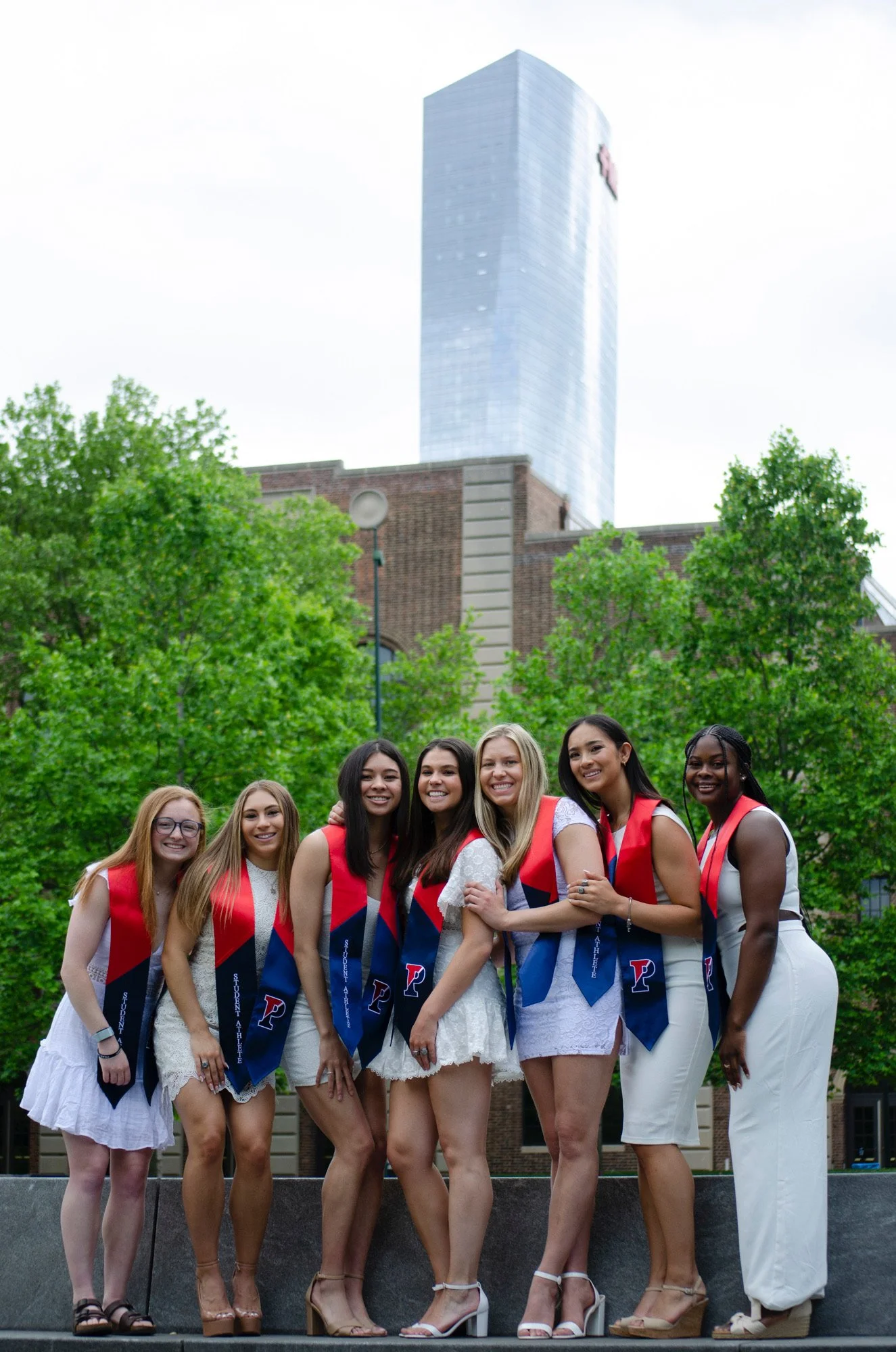 Group of young women in white dresses and red sashes, smiling and posing together outdoors with green trees and a tall modern building in the background.