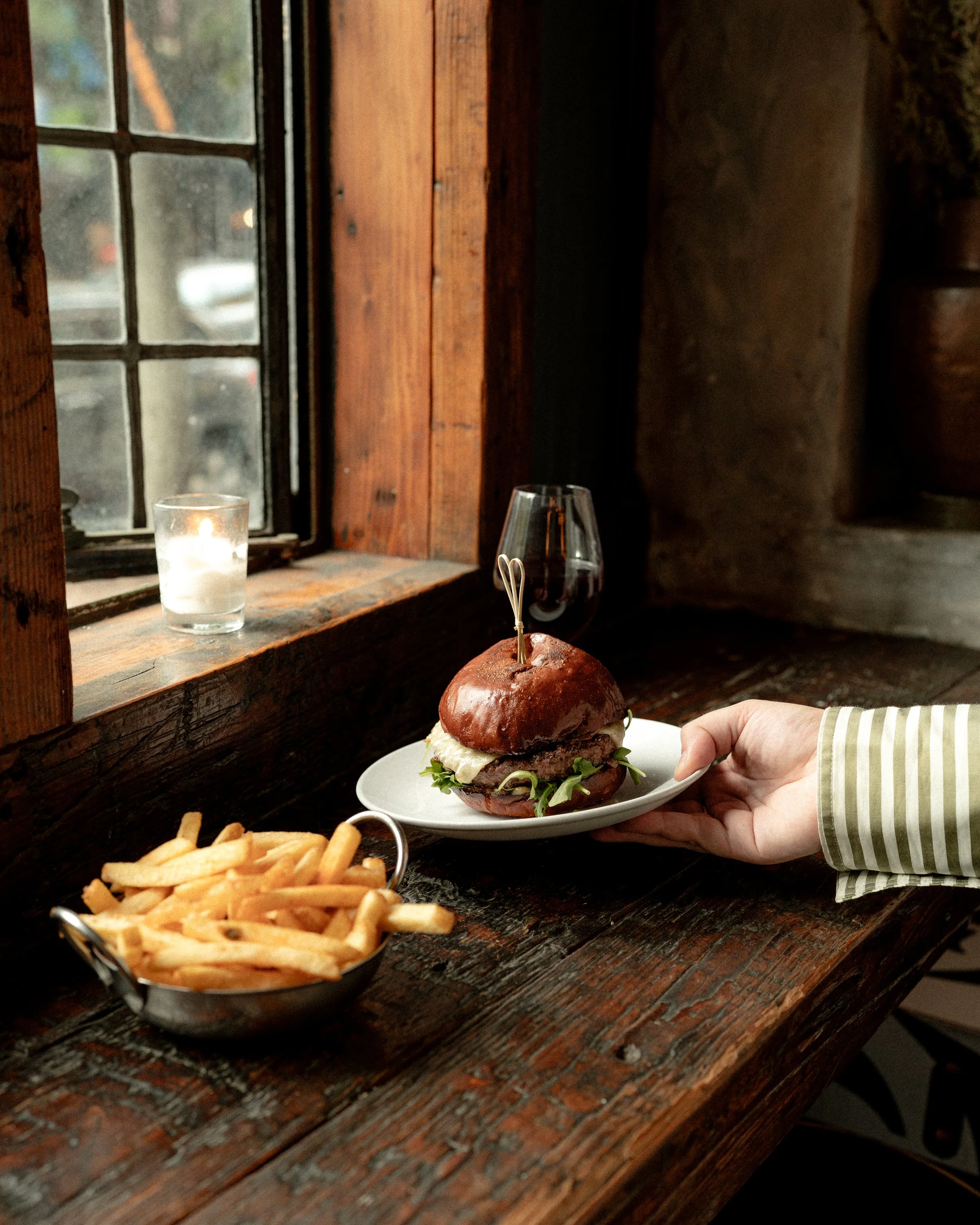 A person is holding a gourmet burger with lettuce, melted cheese, and a beef patty on a white plate. There is also a side of French fries in a metal basket and a glass of red wine on a rustic wooden table near a window with a lit candle.