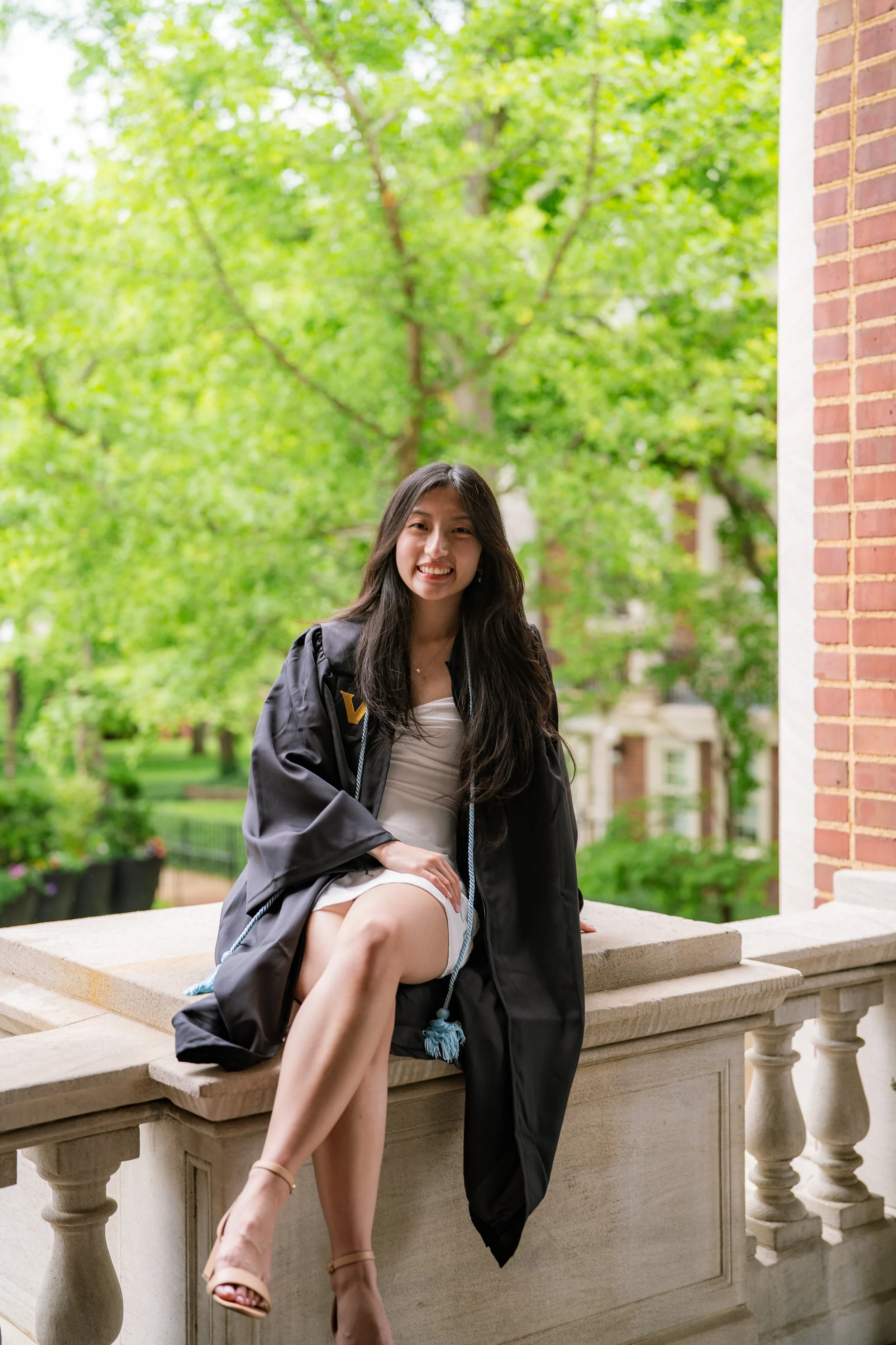 A young woman in a graduation cap with tassels and gown, sitting on a stone balustrade outdoors with green trees in the background.