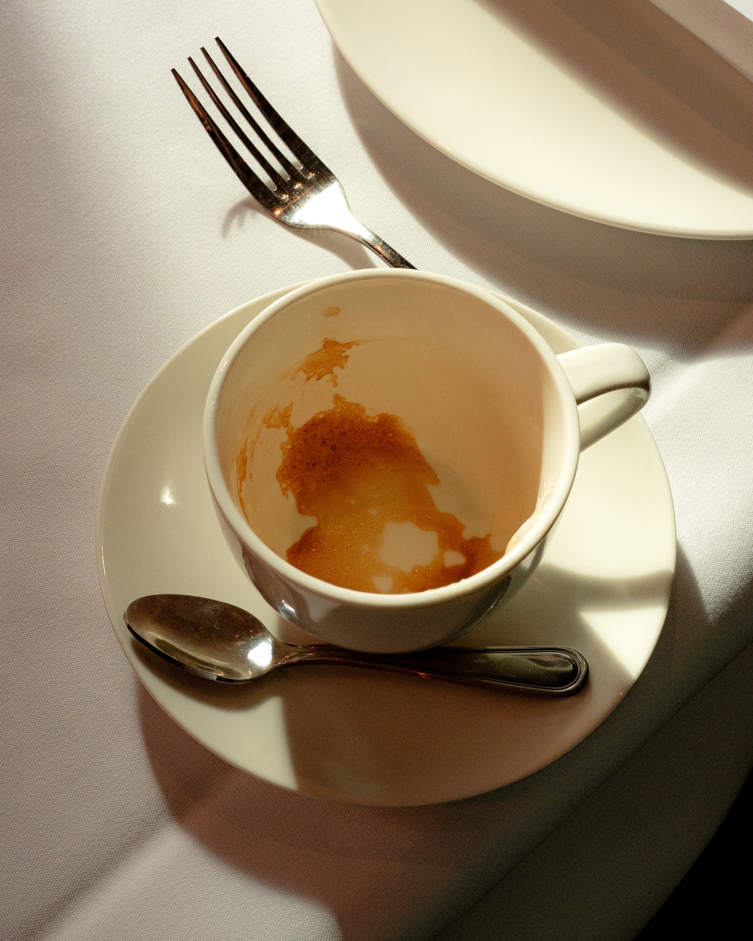 Empty white coffee cup on a saucer with a silver spoon, placed on a white tablecloth. There are some remnants of a beverage inside the cup.