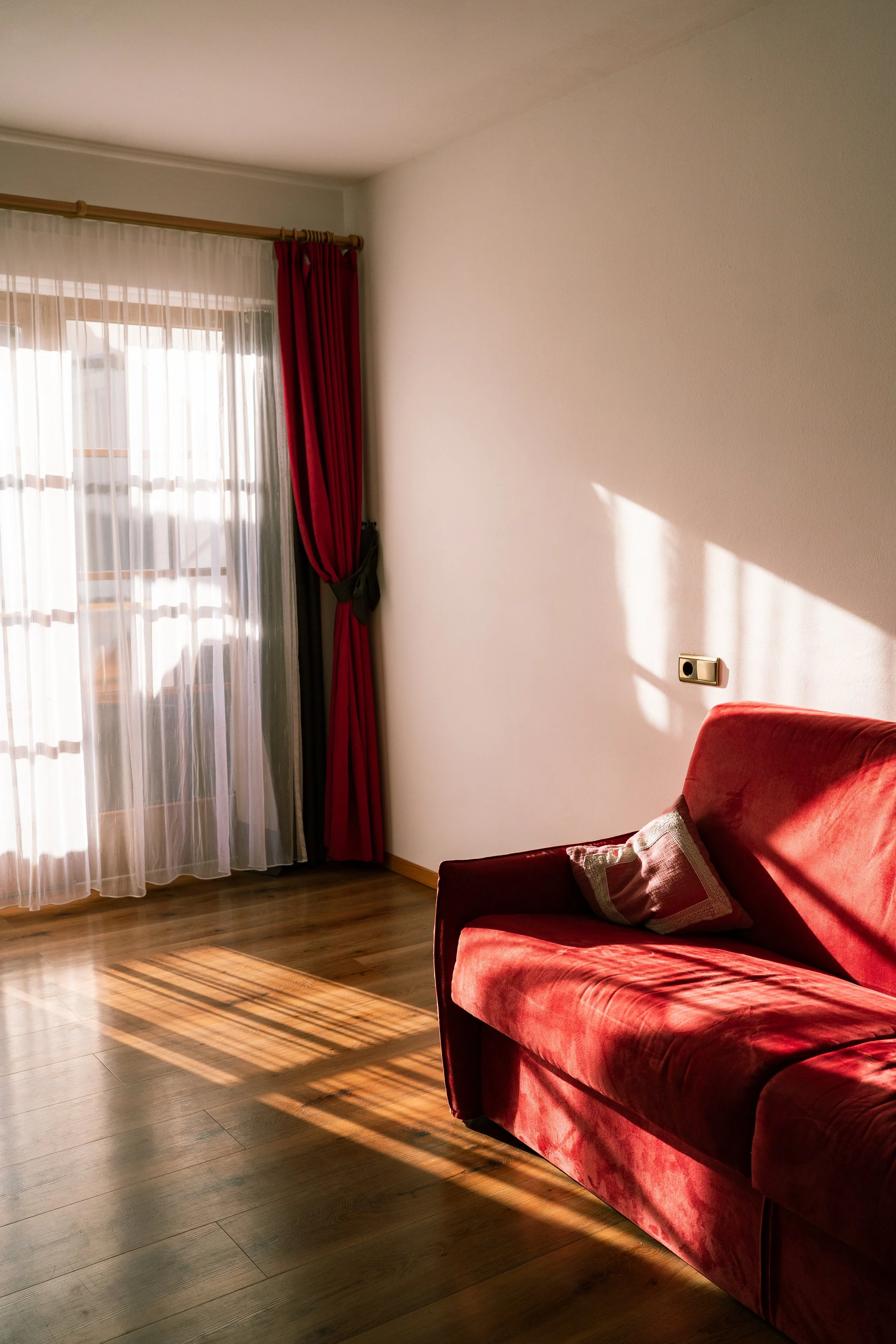 Sunlight streaming through a window with sheer and red curtains, casting shadows on a wooden floor next to a red velvet sofa with a pillow.