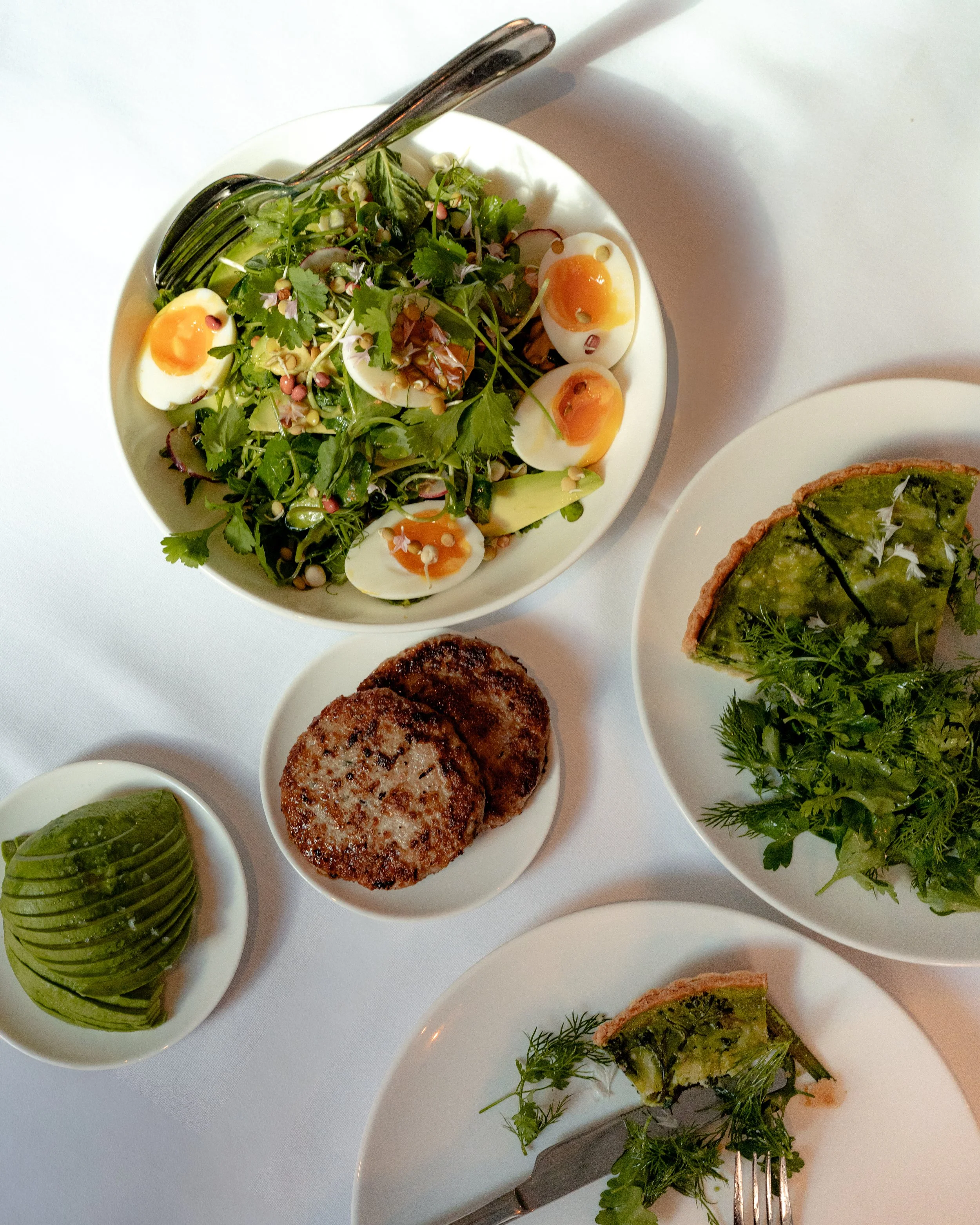 A white bowl contains a mixed greens salad with boiled eggs, microgreens, and sprouts. There is a transparent serving spoon resting on the bowl. To the right, there is a piece of quiche with a green vegetable filling garnished with herbs. Below, a sm