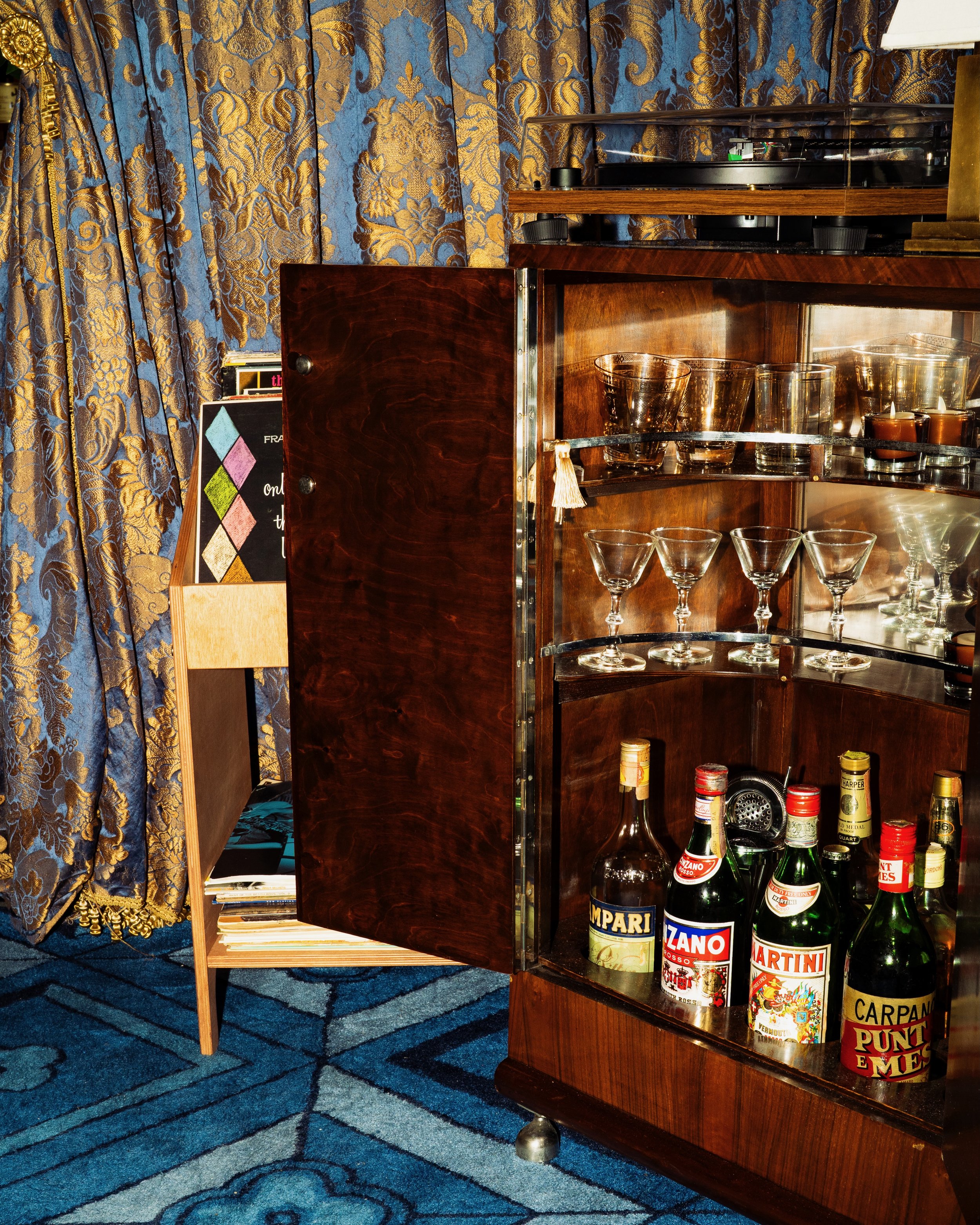 A wooden bar cabinet with a glass top, containing several bottles of alcohol with colorful labels, glasses, and candles. Blue and gold patterned curtains are seen in the background.