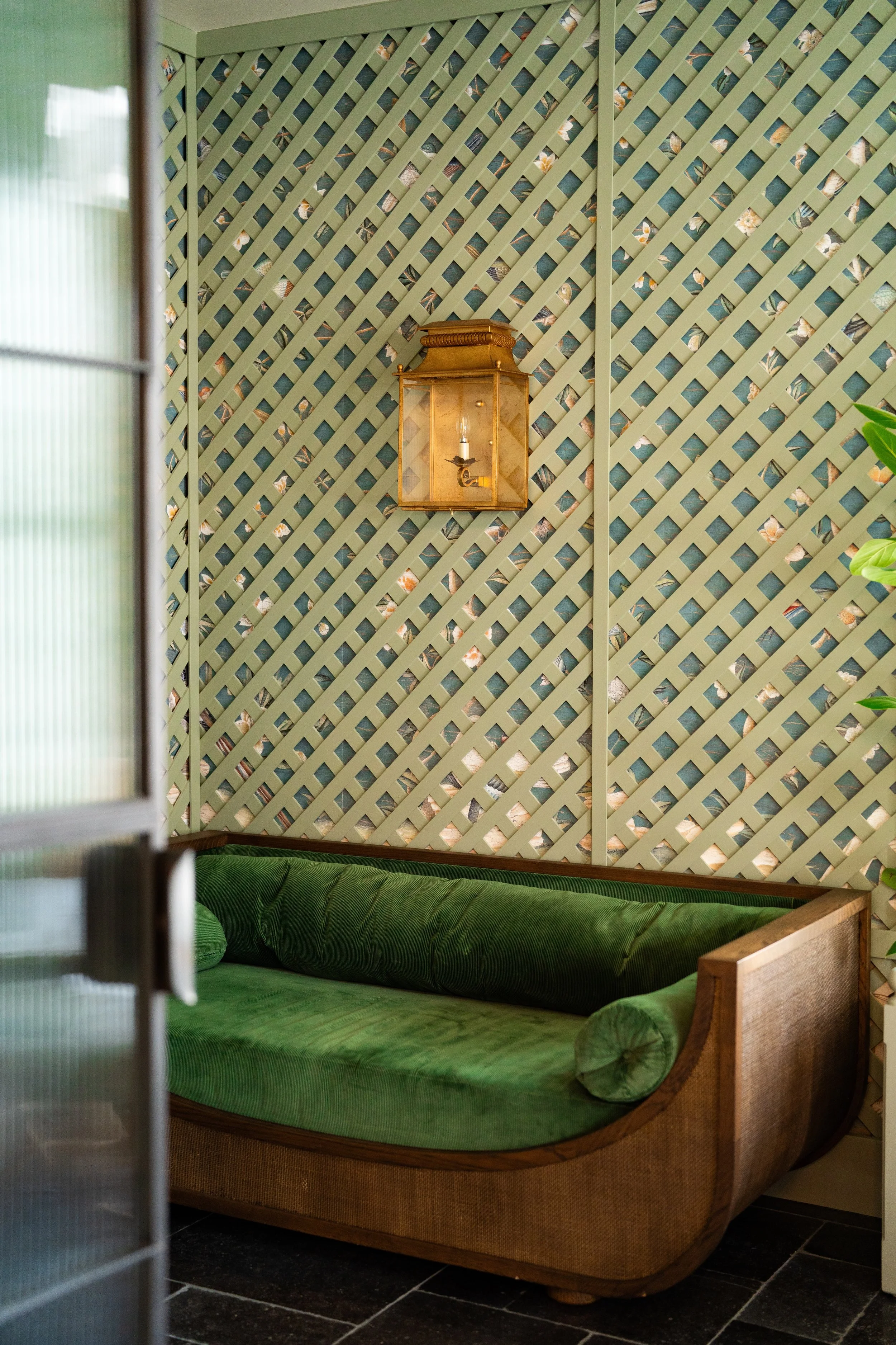 A vintage green velvet sofa with a curved wooden frame and a single cylindrical pillow, situated in front of a decorative wallpapered wall with a geometric pattern and a wall-mounted brass and glass lantern light fixture.