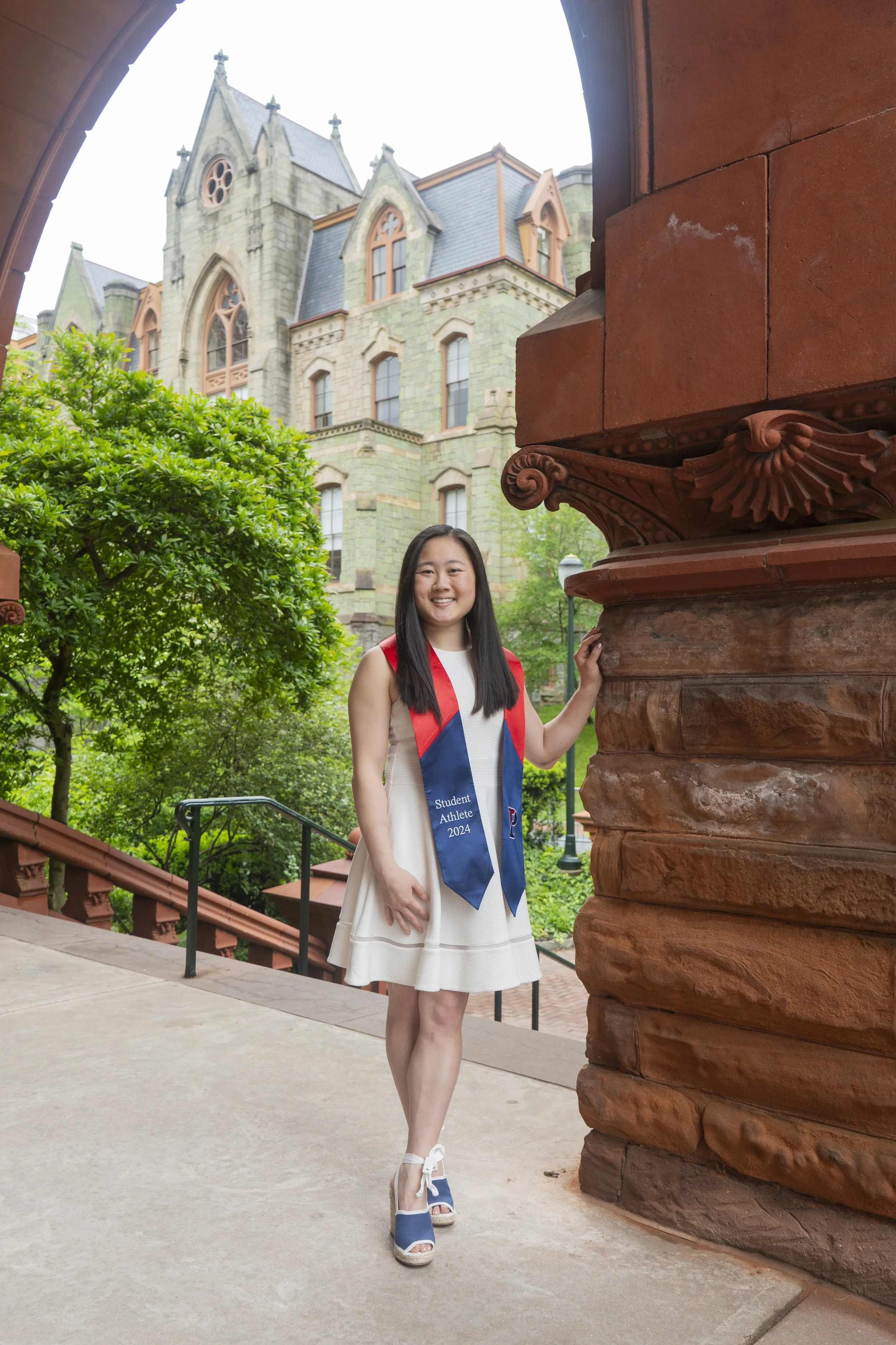 A young woman in a white dress and blue high-heeled shoes stands on a sidewalk, holding a red and blue graduation stole that reads 'Student Athlete 2024.' She smiles at the camera, with a historic stone building with pointed windows and a tree with l