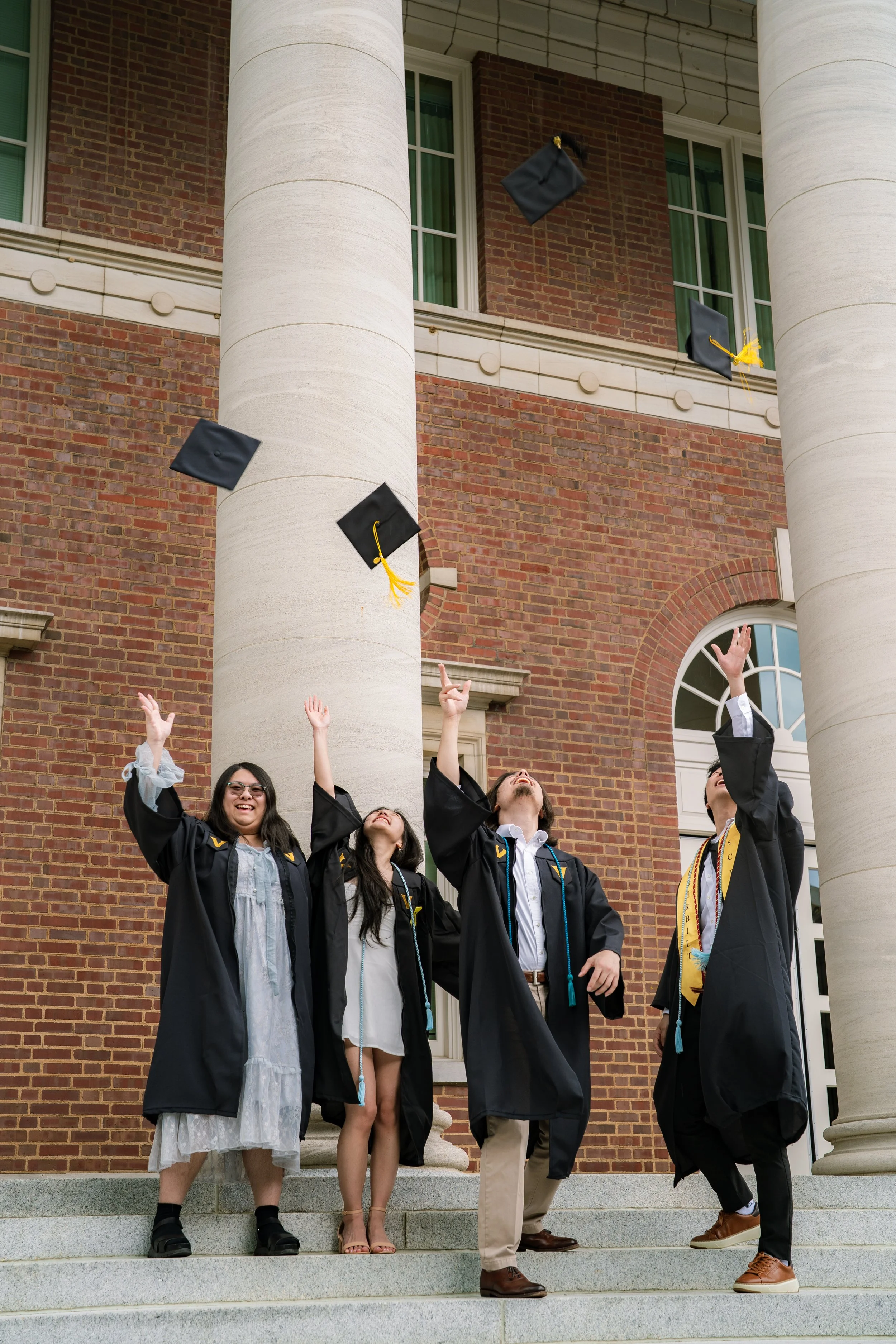 Group of diverse graduates in caps and gowns celebrating graduation outside a brick building with large columns, tossing caps into the air.