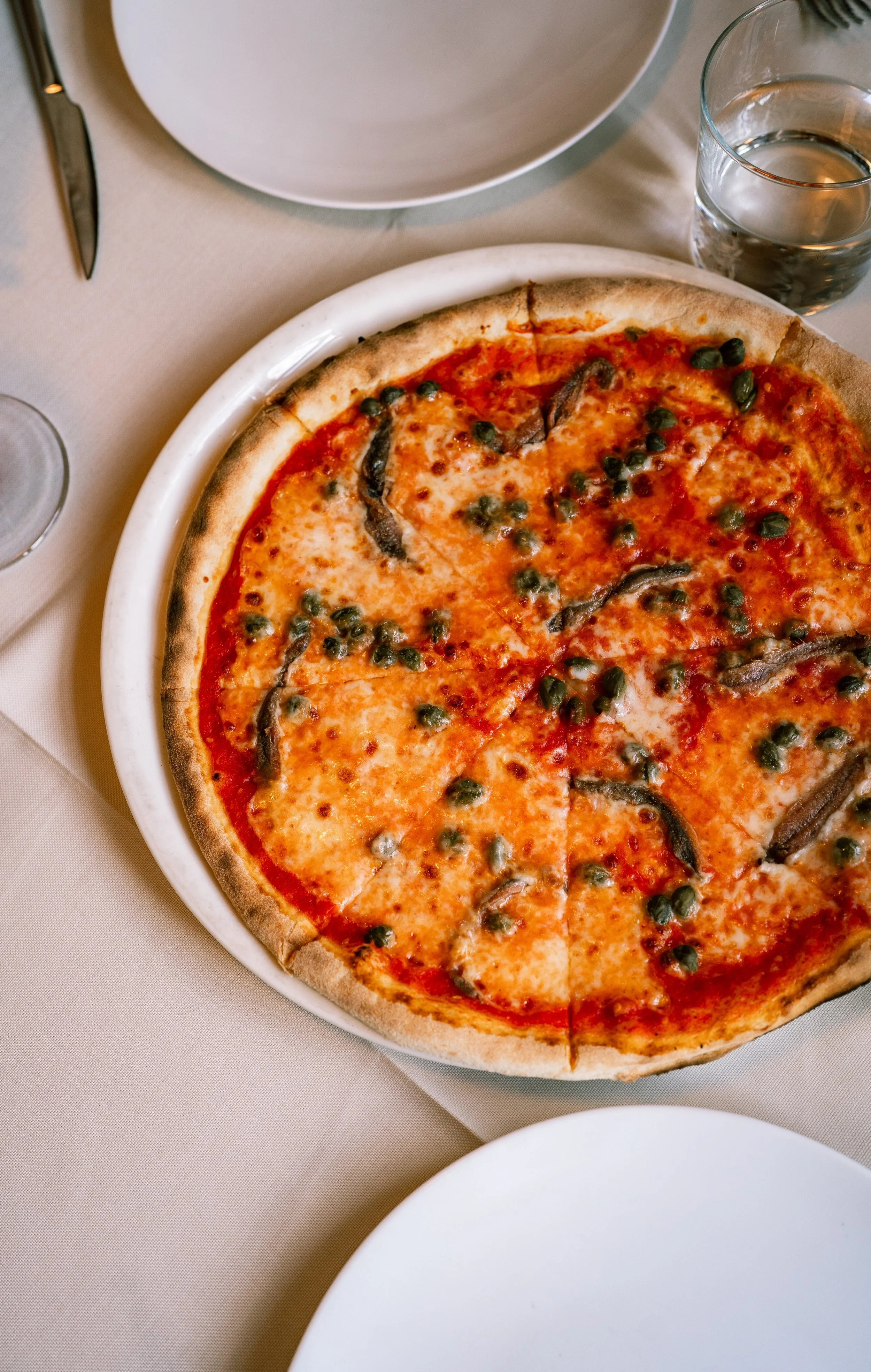 A pizza with cheese, pepperoni, green peppers, and black olives in a white ceramic dish, set on a beige tablecloth.