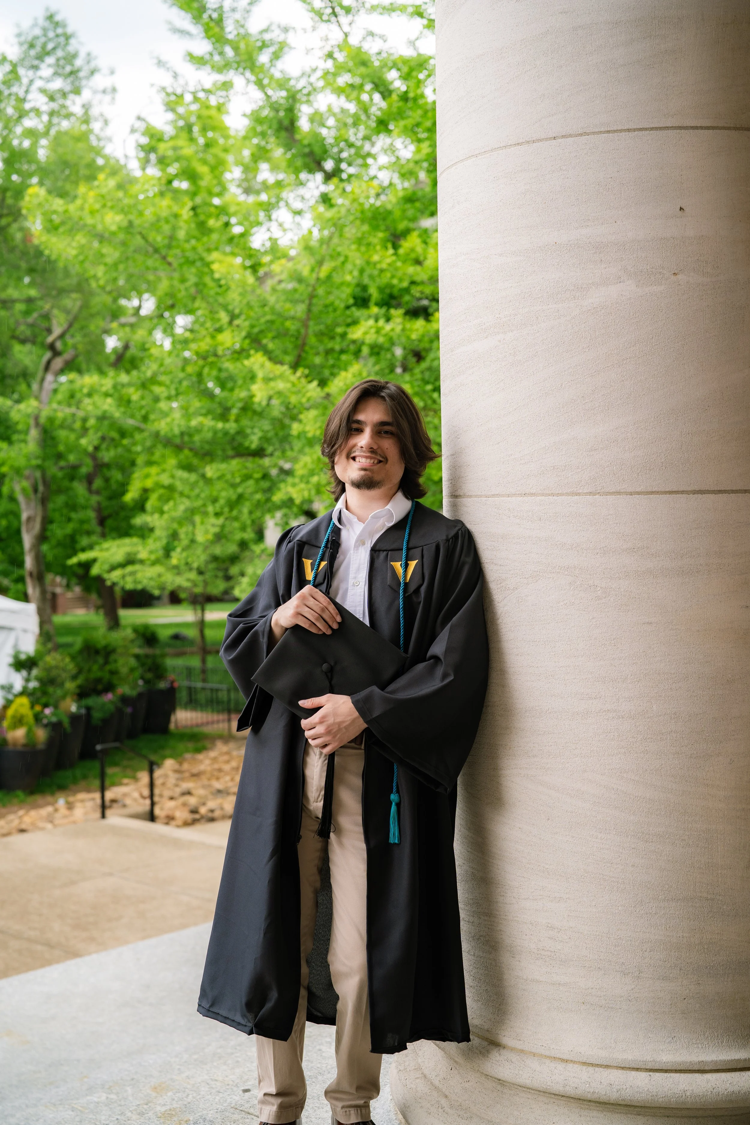 Young man in graduation cap and gown leaning against a large beige stone column outside on a university campus, with green trees in the background.