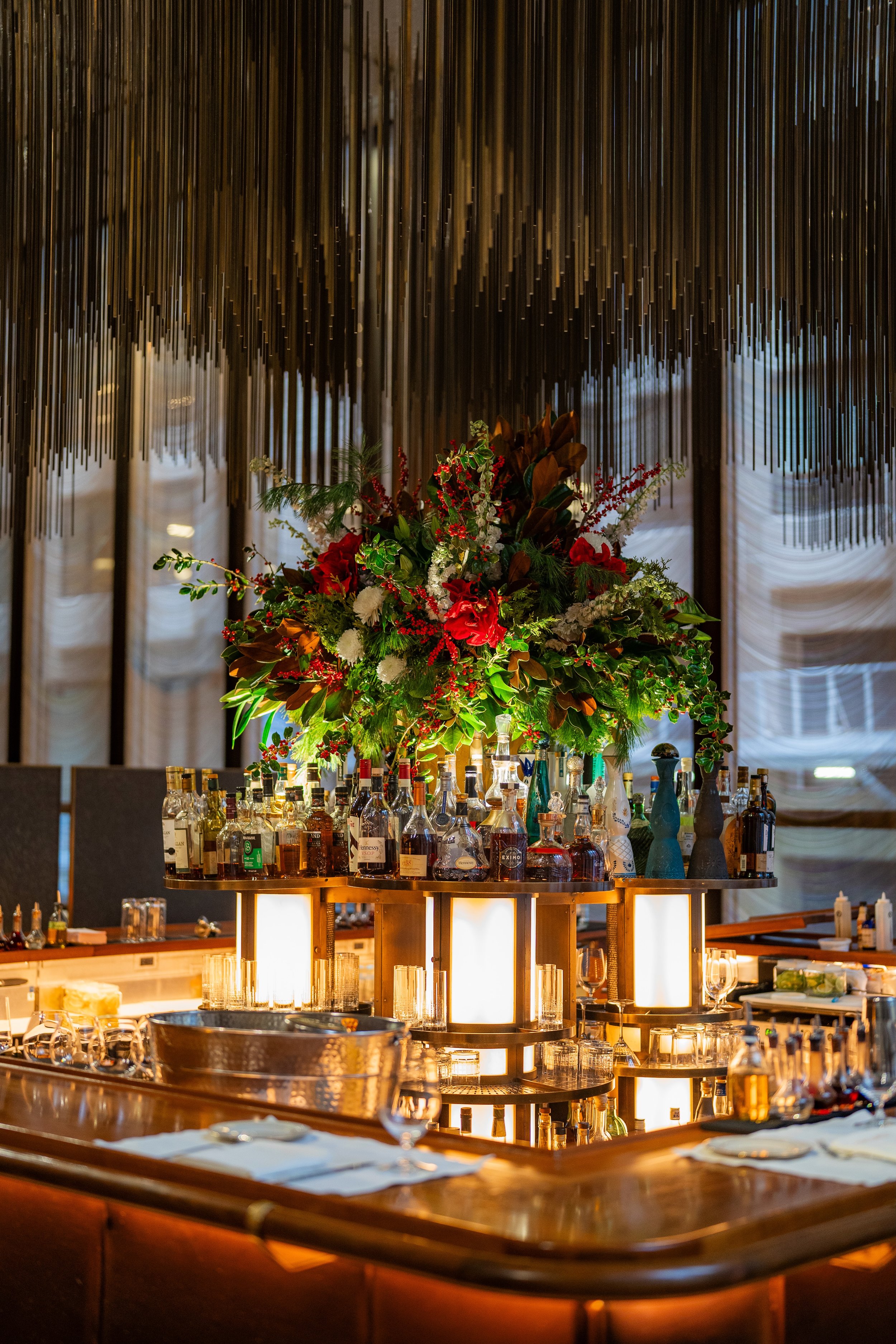 A bar with a large floral arrangement and bottles of liquor. The background features decorative hanging metal rods and draped curtains.
