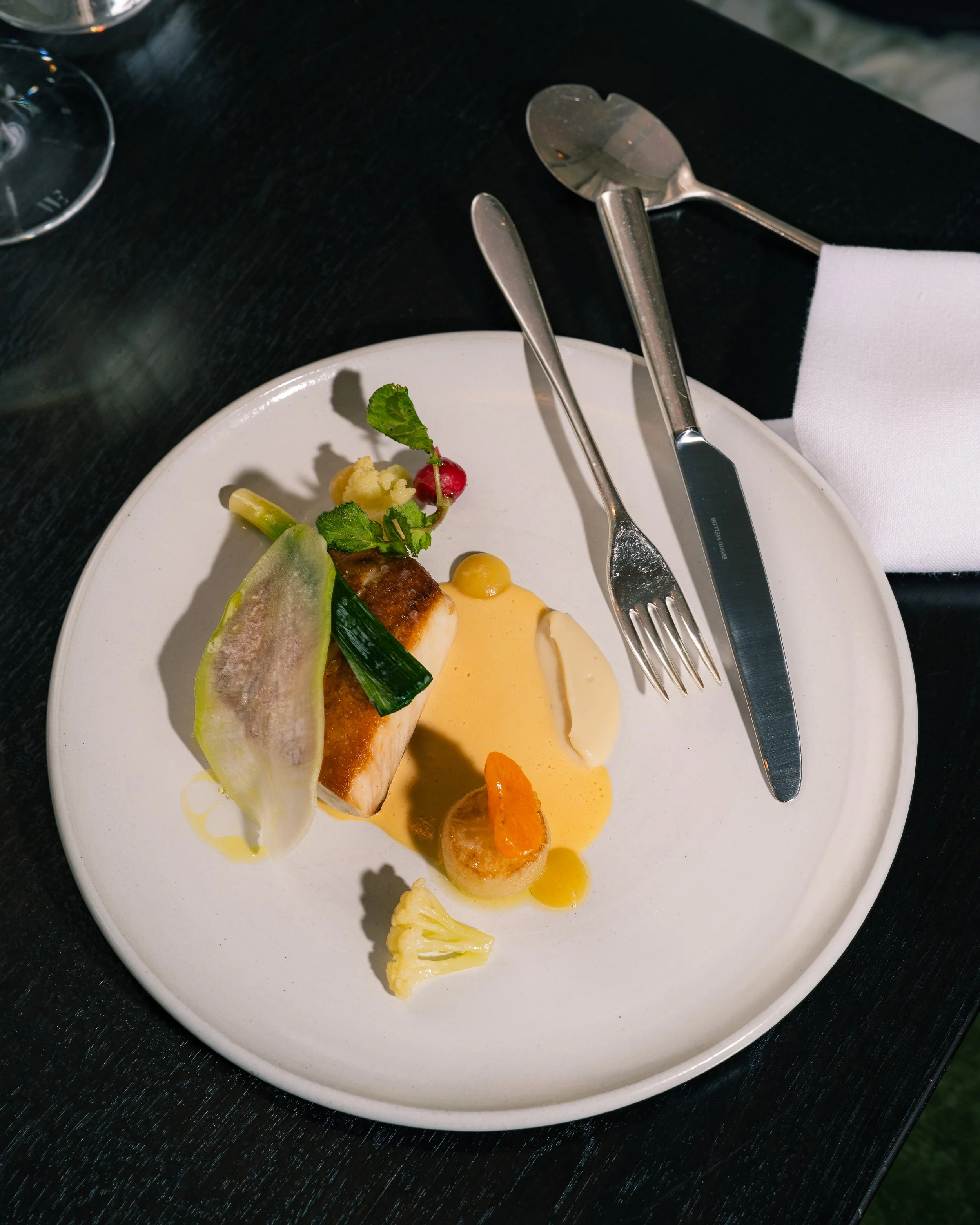 A plated dish with white fish, green vegetables, radish, and a yellow sauce on a white plate, with a steak knife and fork, on a black table.