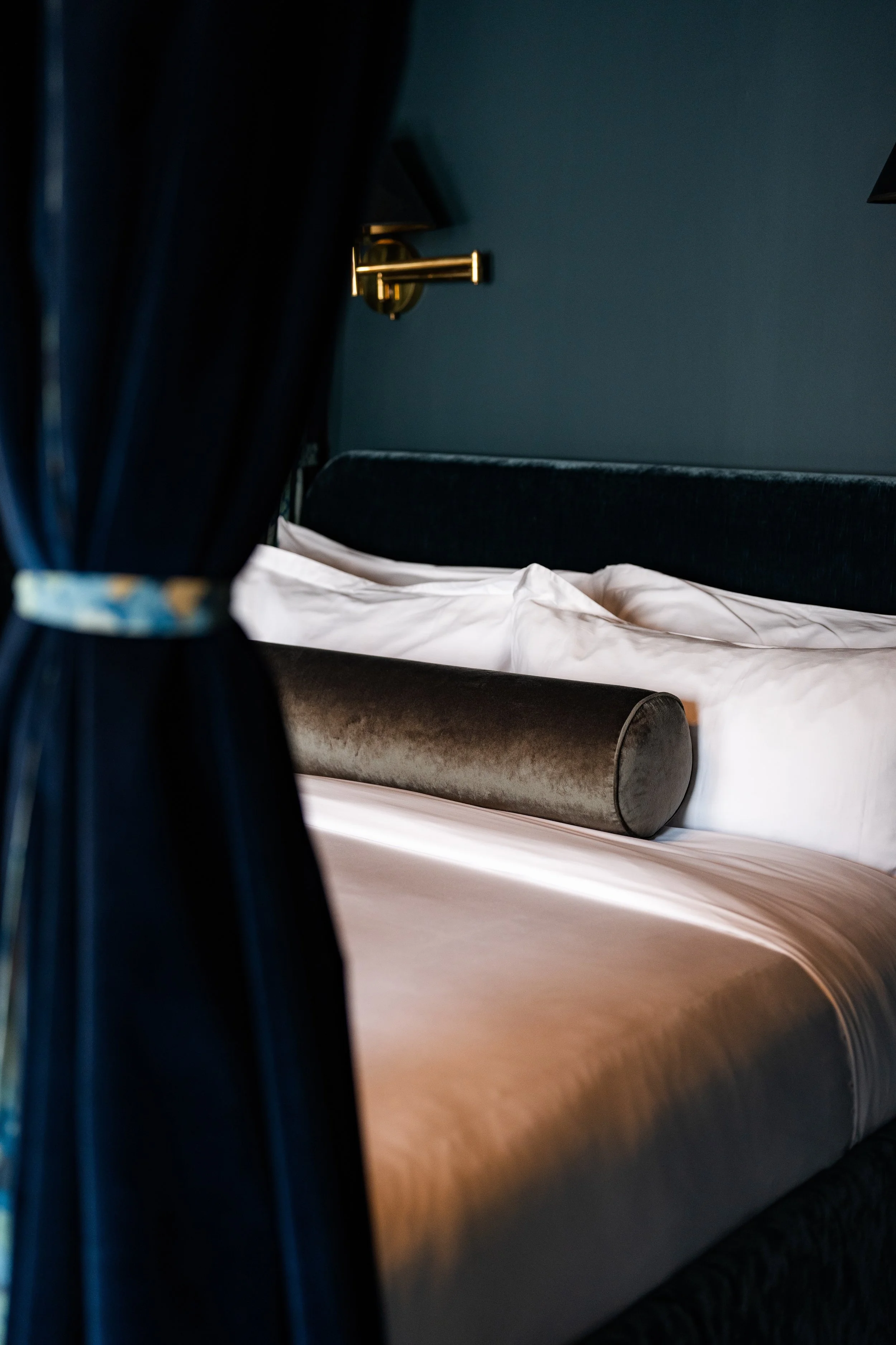 A bed with a white sheet, a dark headboard, a velvet pillow, and a brass wall lamp on a dark blue wall.