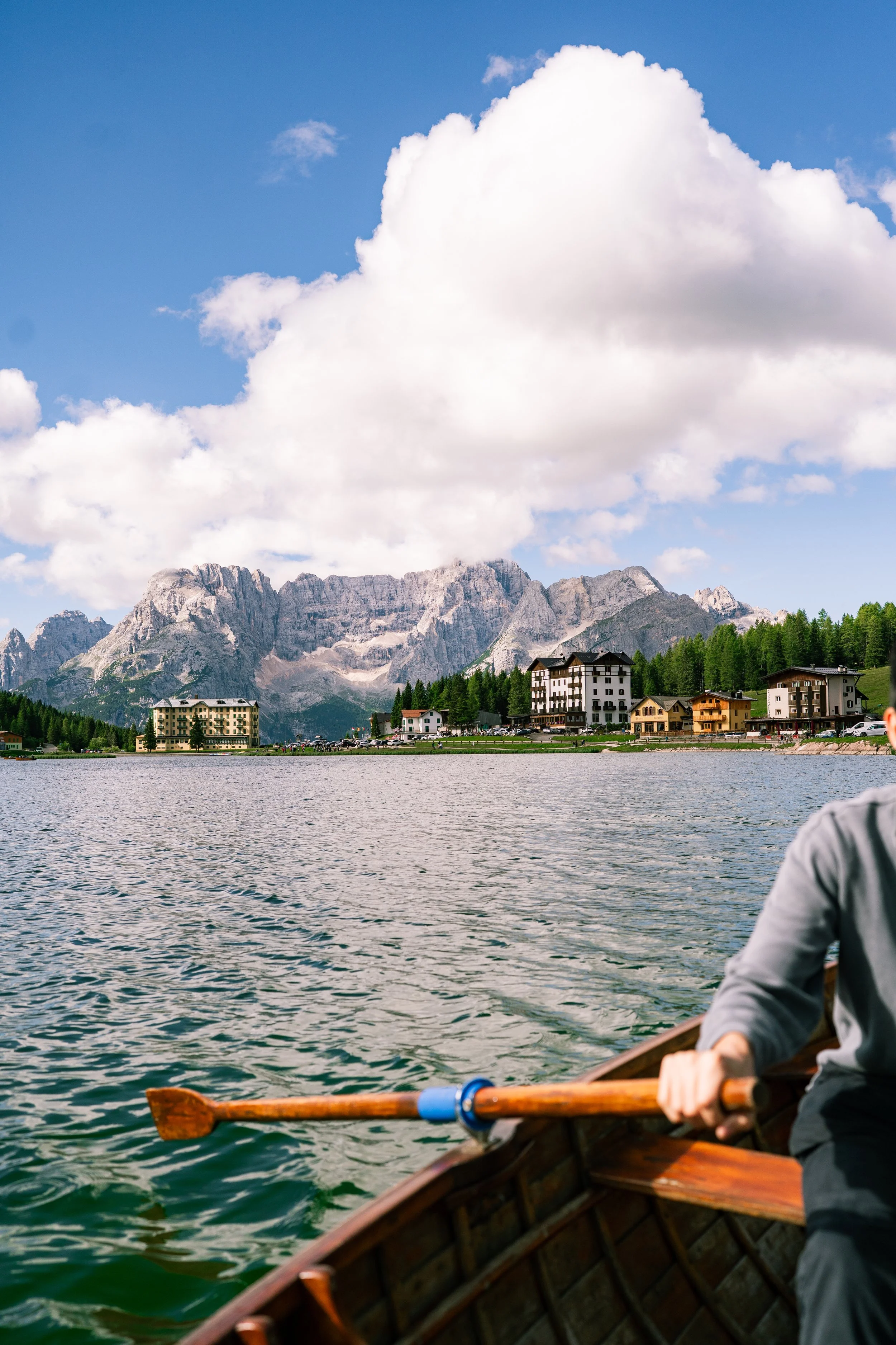 A person rowing a boat on a lake with mountains and buildings in the background, under a partly cloudy sky.