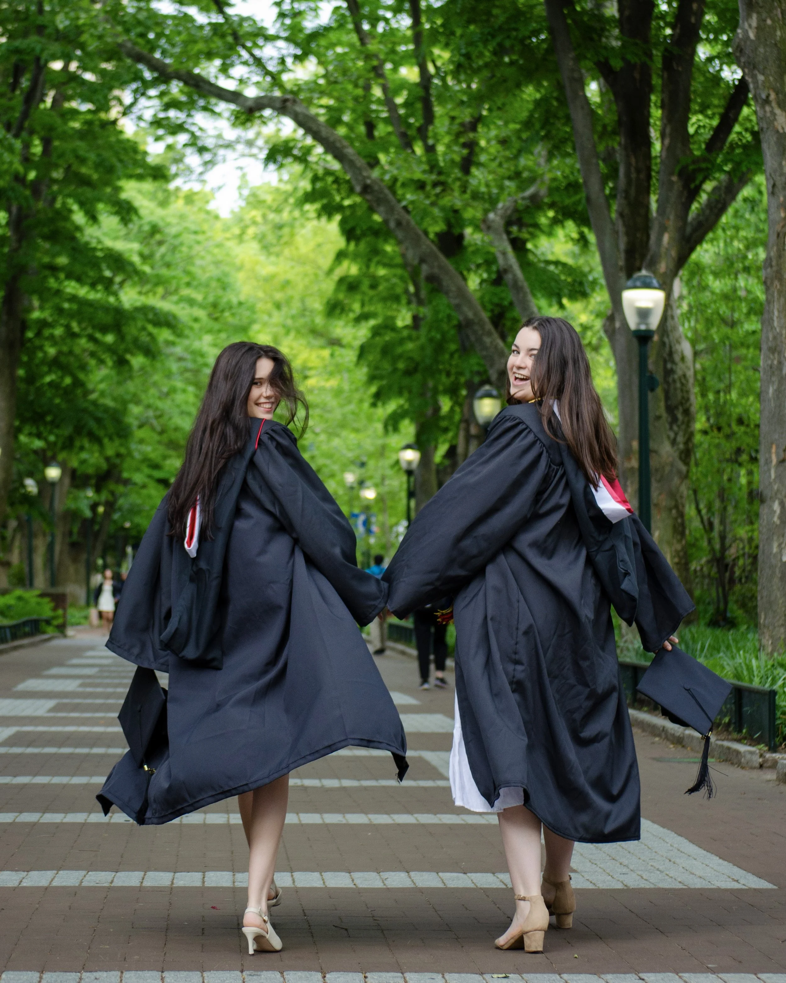 Two young women in graduation gowns holding hands, walking away from camera in a park with green trees and lampposts.