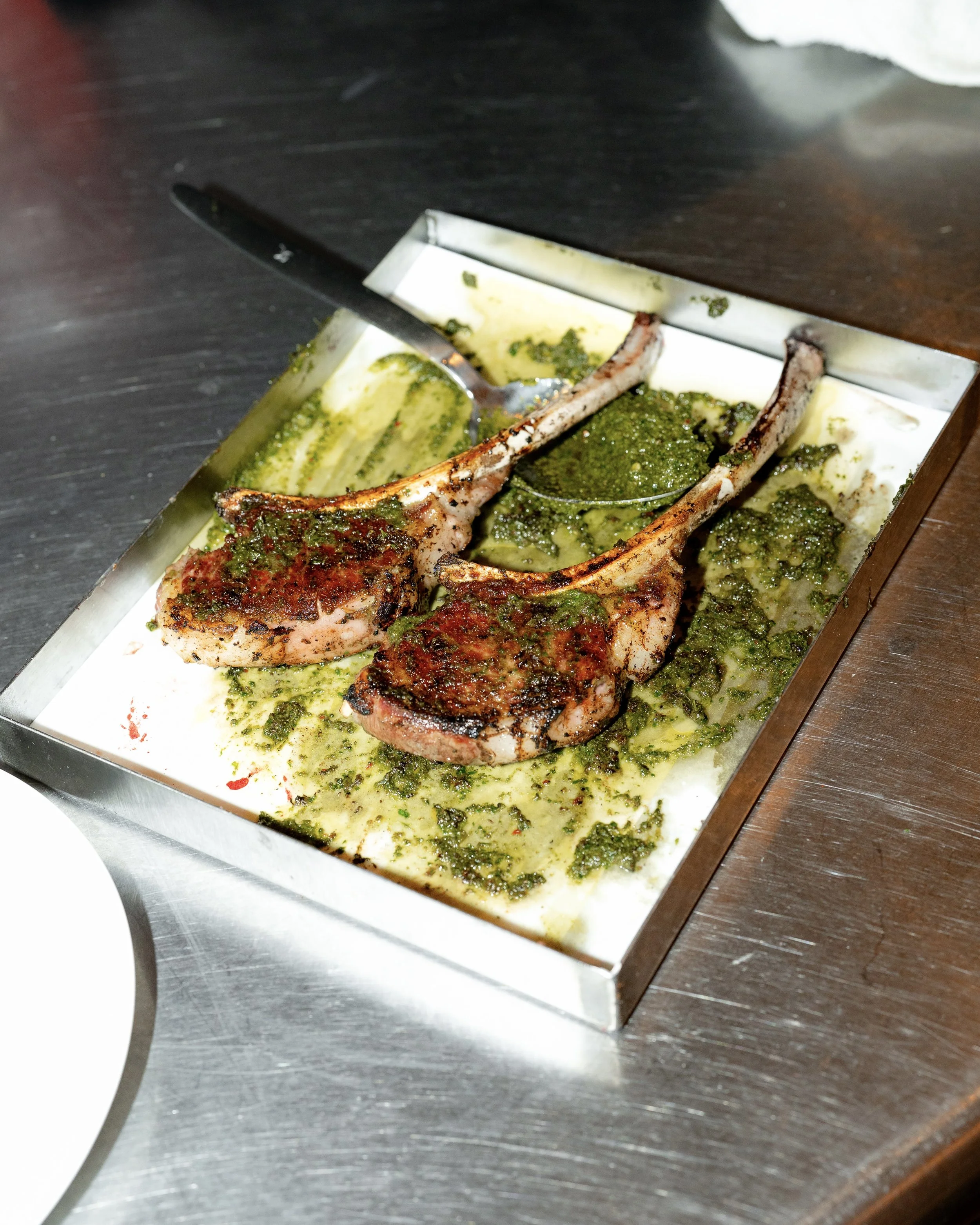 A pair of grilled lamb chops topped with green herb sauce on a stainless steel tray.
