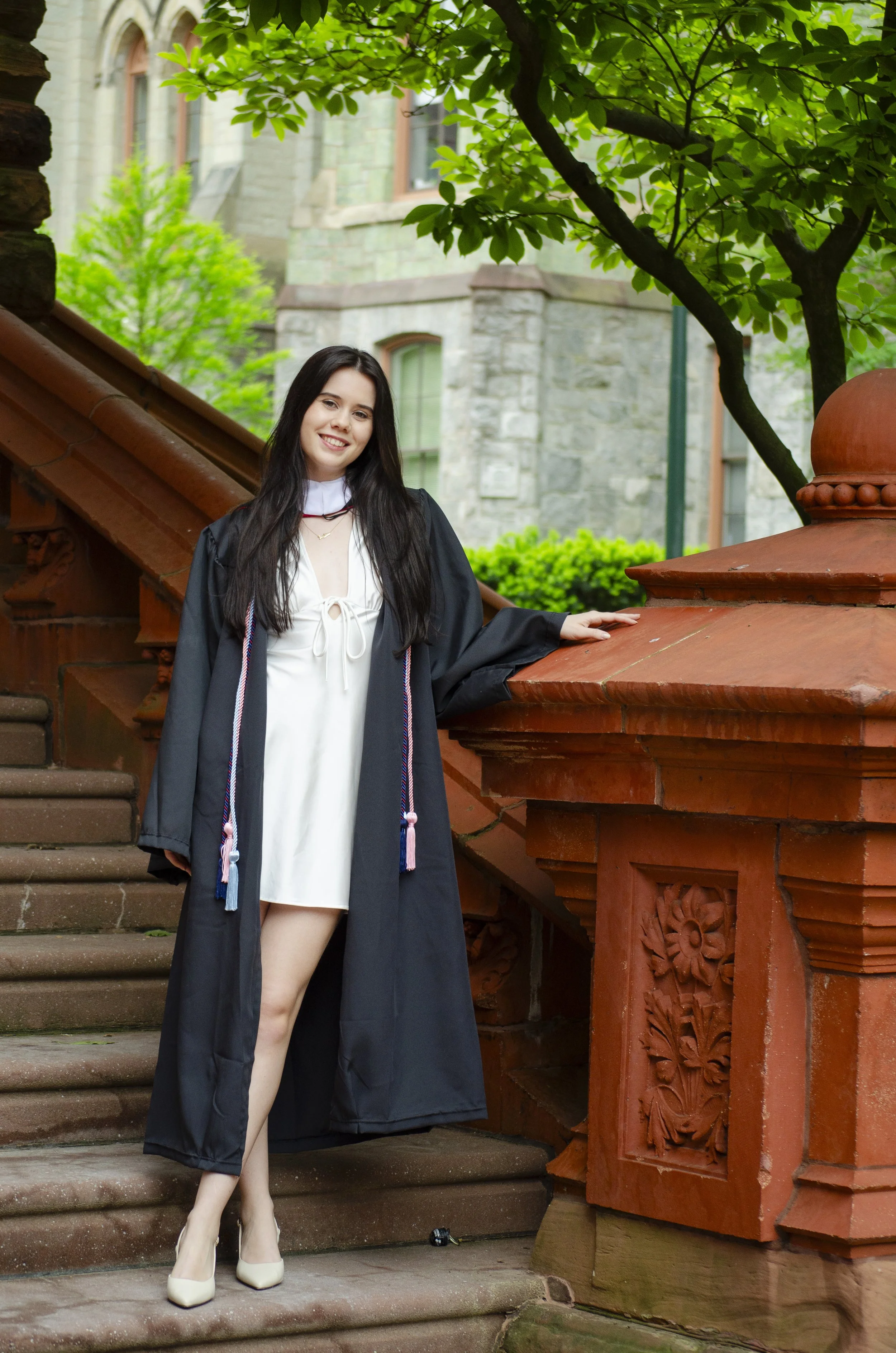 Young woman in graduation cap and gown standing on stone steps in front of a historic building with trees.